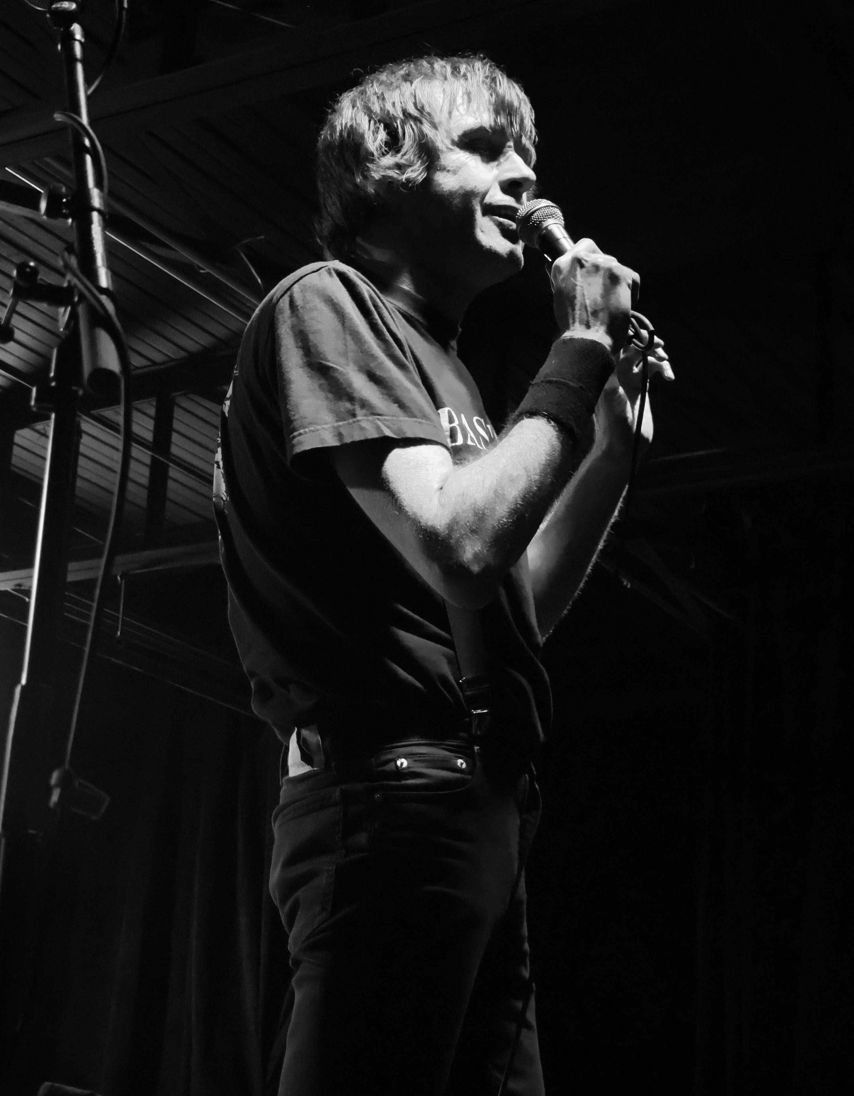 A performer singing into a microphone on stage, dressed in a black t-shirt and dark jeans, captured in black and white with dramatic lighting.