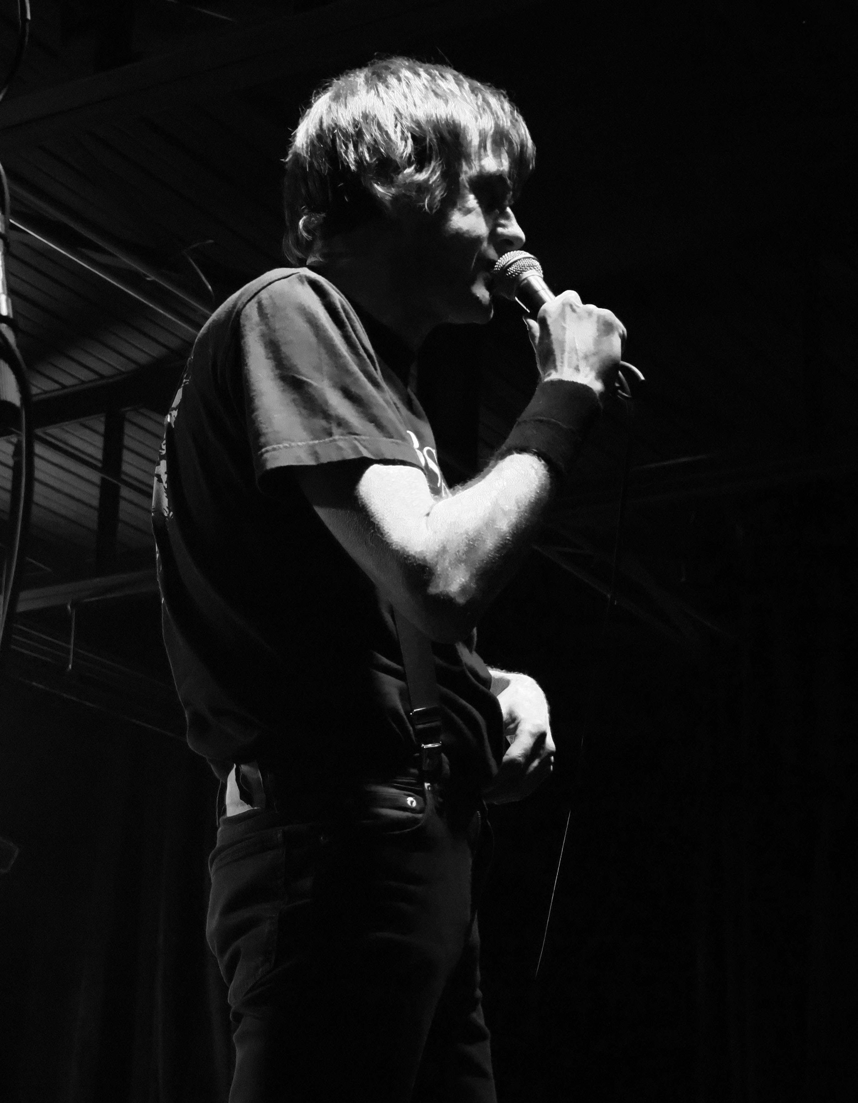 A black and white image of a singer speaking into a microphone on stage, wearing a black t-shirt and dark pants, with a focus on their profile.