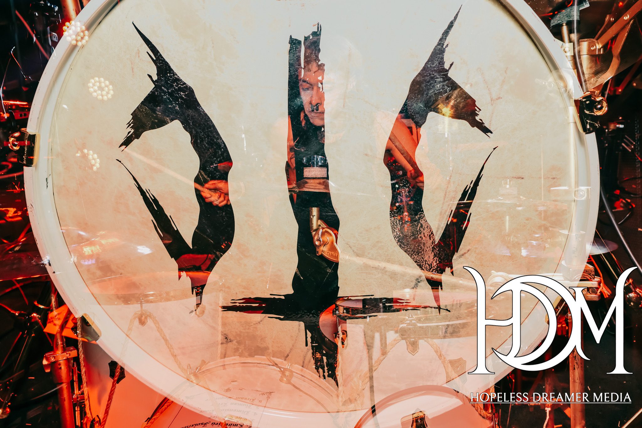 A close-up of a drum kit featuring a drumhead with an artistic black design and a reflection of a drummer's hands, set against a vibrant, illuminated stage background.