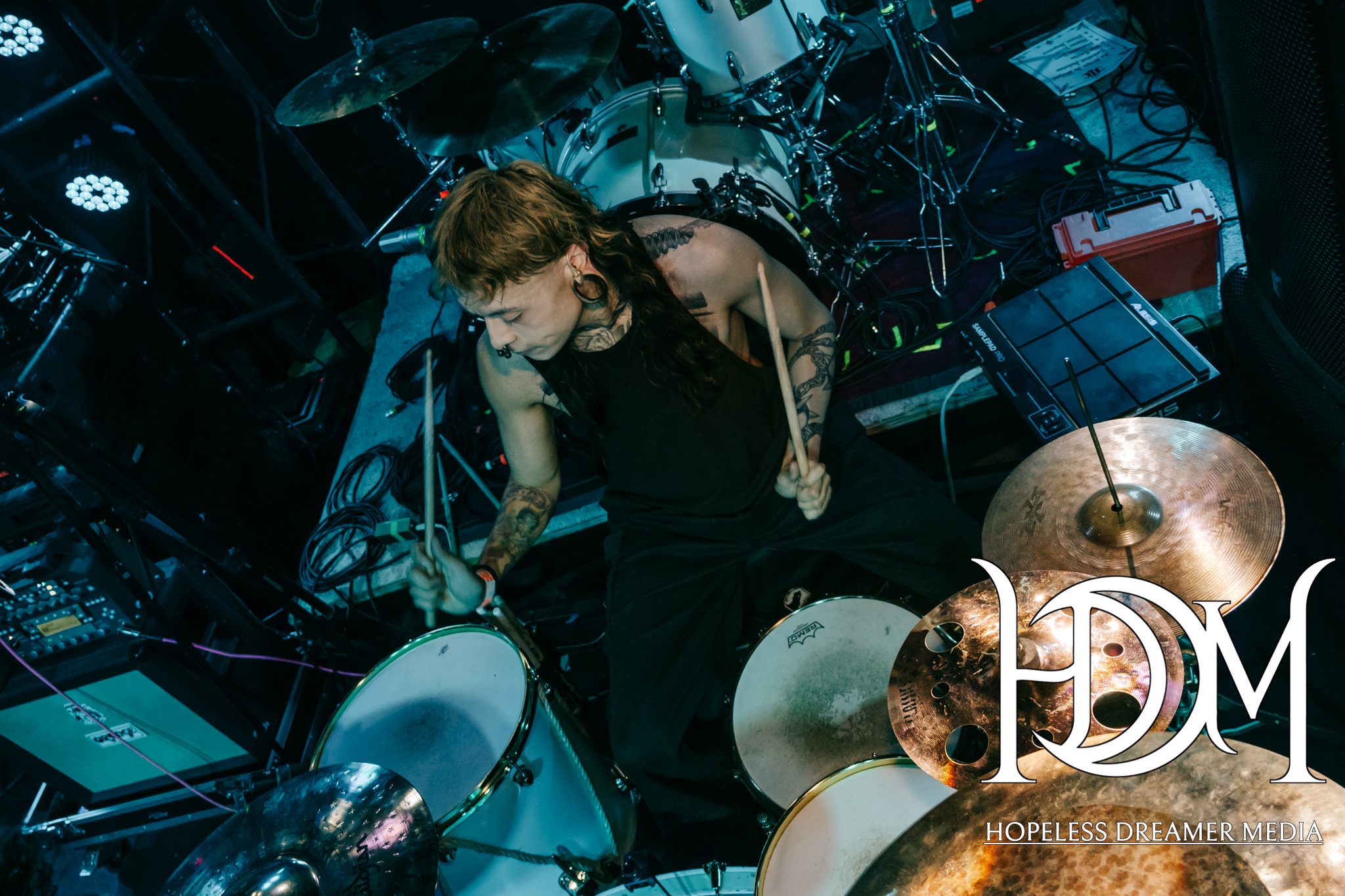 A drummer performing energetically on stage, surrounded by a drum kit and other music equipment.