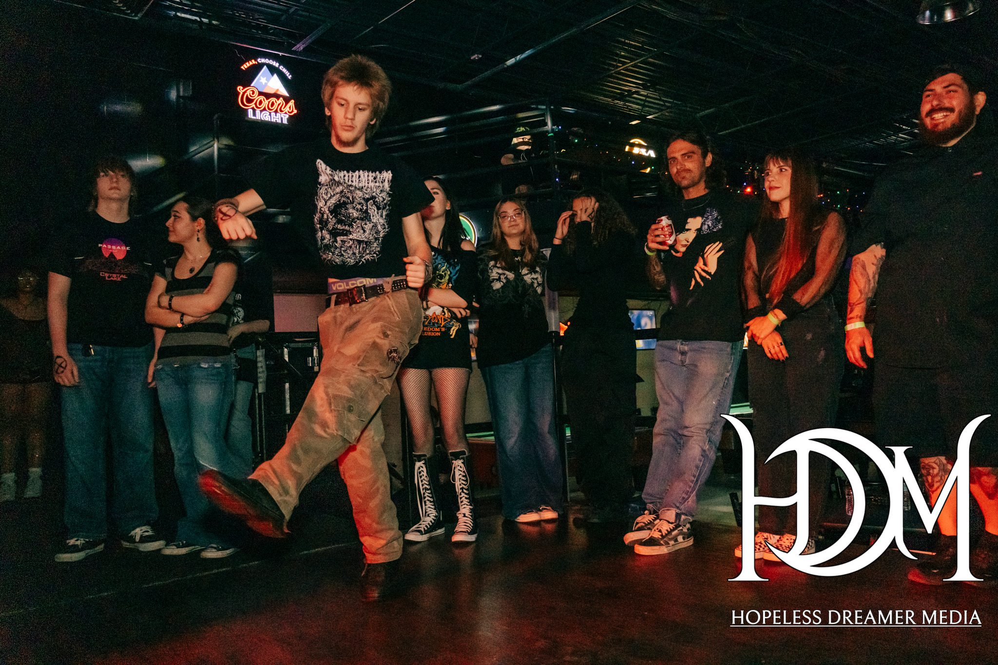 A group of young people enjoying a live music event at The Zone Bar in Abilene, TX. One person is energetically dancing in the foreground, while others watch and engage in conversation. The setting includes a neon Coors Light sign in the background, reflecting a vibrant atmosphere.
