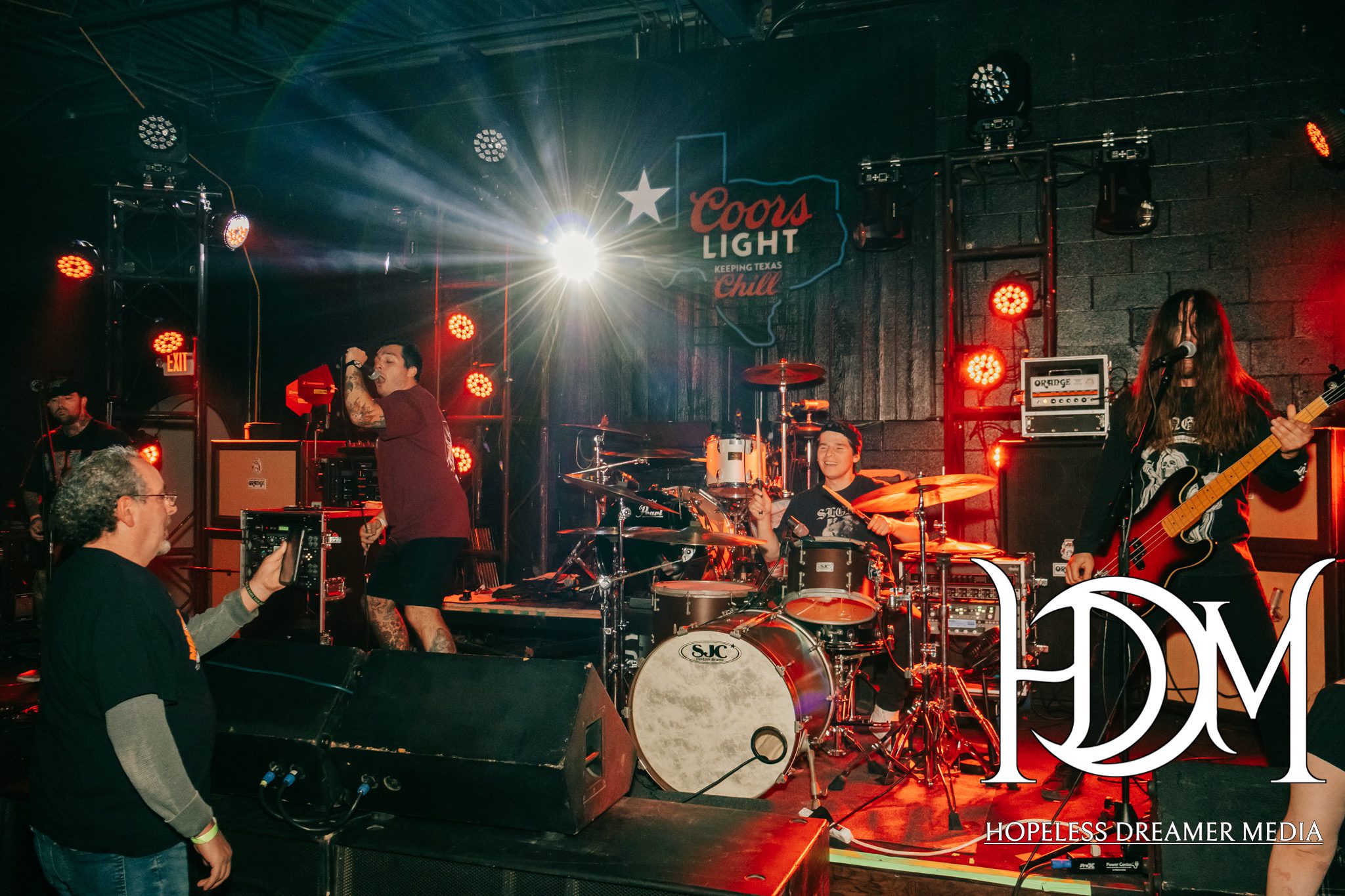 A live performance by a metalcore band on stage, featuring the vocalist engaging with the audience, a drummer playing energetically, and a bassist contributing to the sound, with dramatic lighting effects and a music venue backdrop.