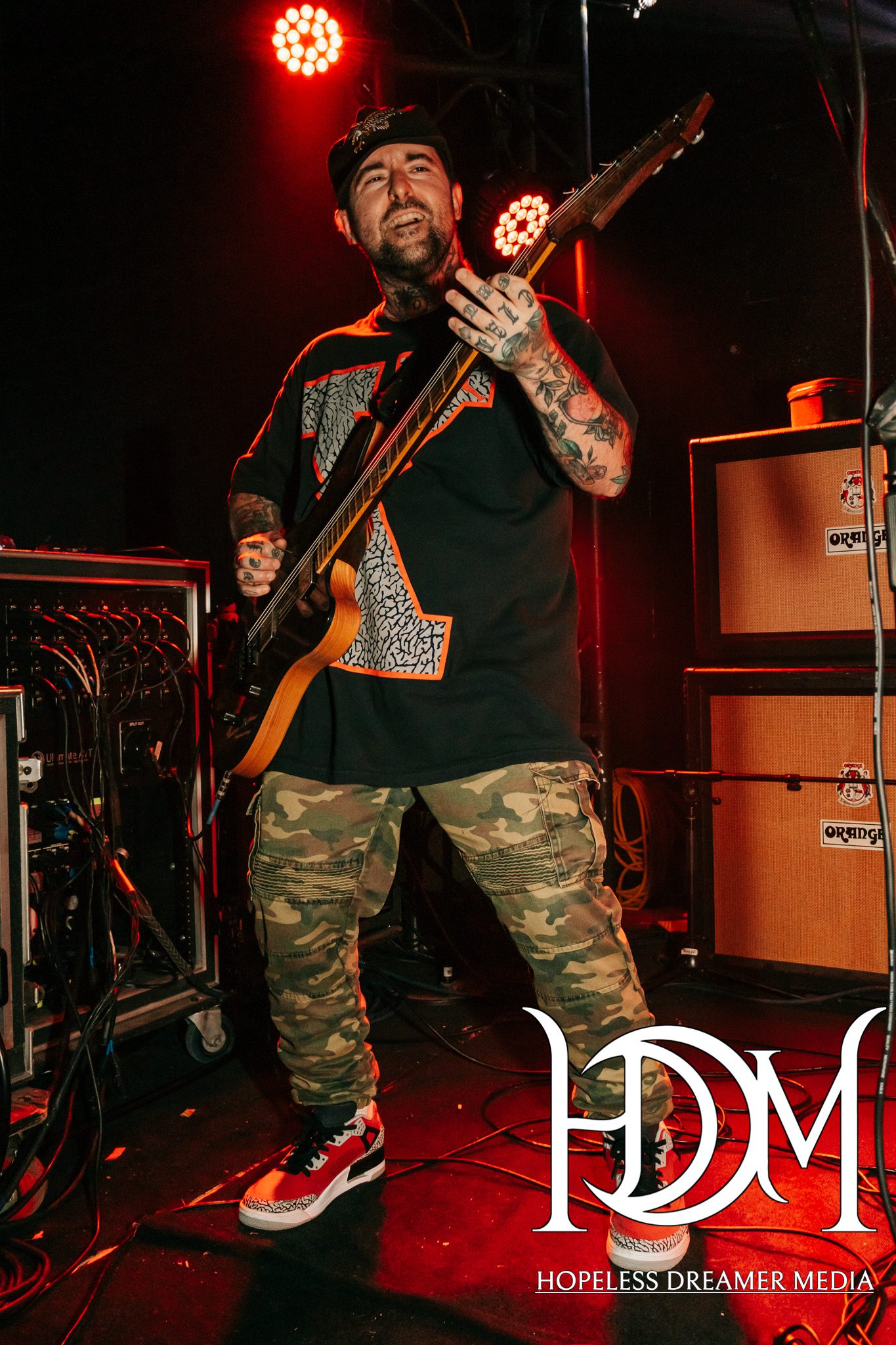 A guitarist performing on stage, wearing a black shirt with an orange design and camouflage pants, smiling while playing an electric guitar, with bright red stage lights in the background.