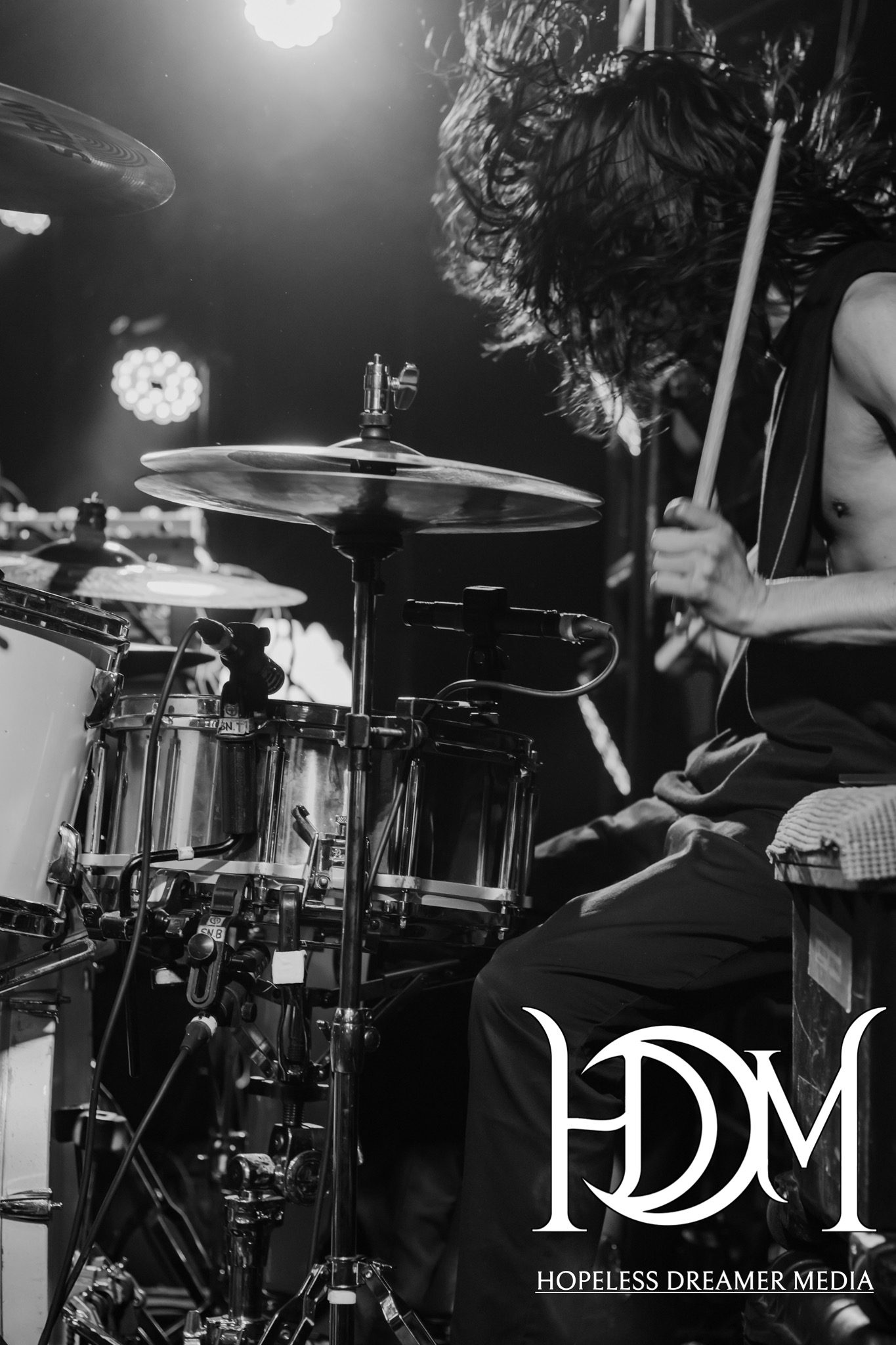 A drummer passionately playing on stage, with intense focus and flowing hair, illuminated by stage lights.