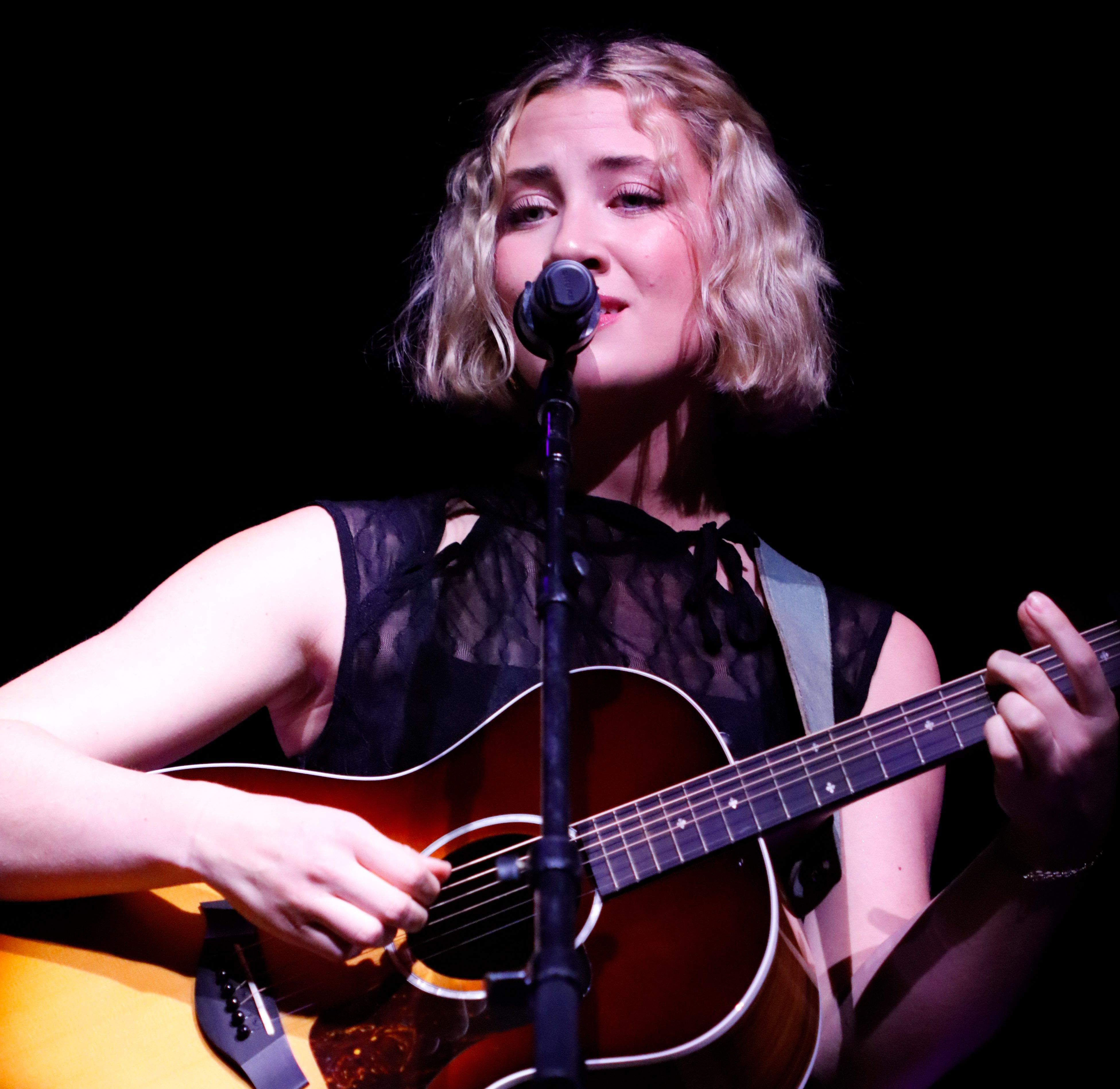 A musician with curly hair sings passionately into a microphone while playing an acoustic guitar on stage, dressed in a stylish black outfit against a dark backdrop.