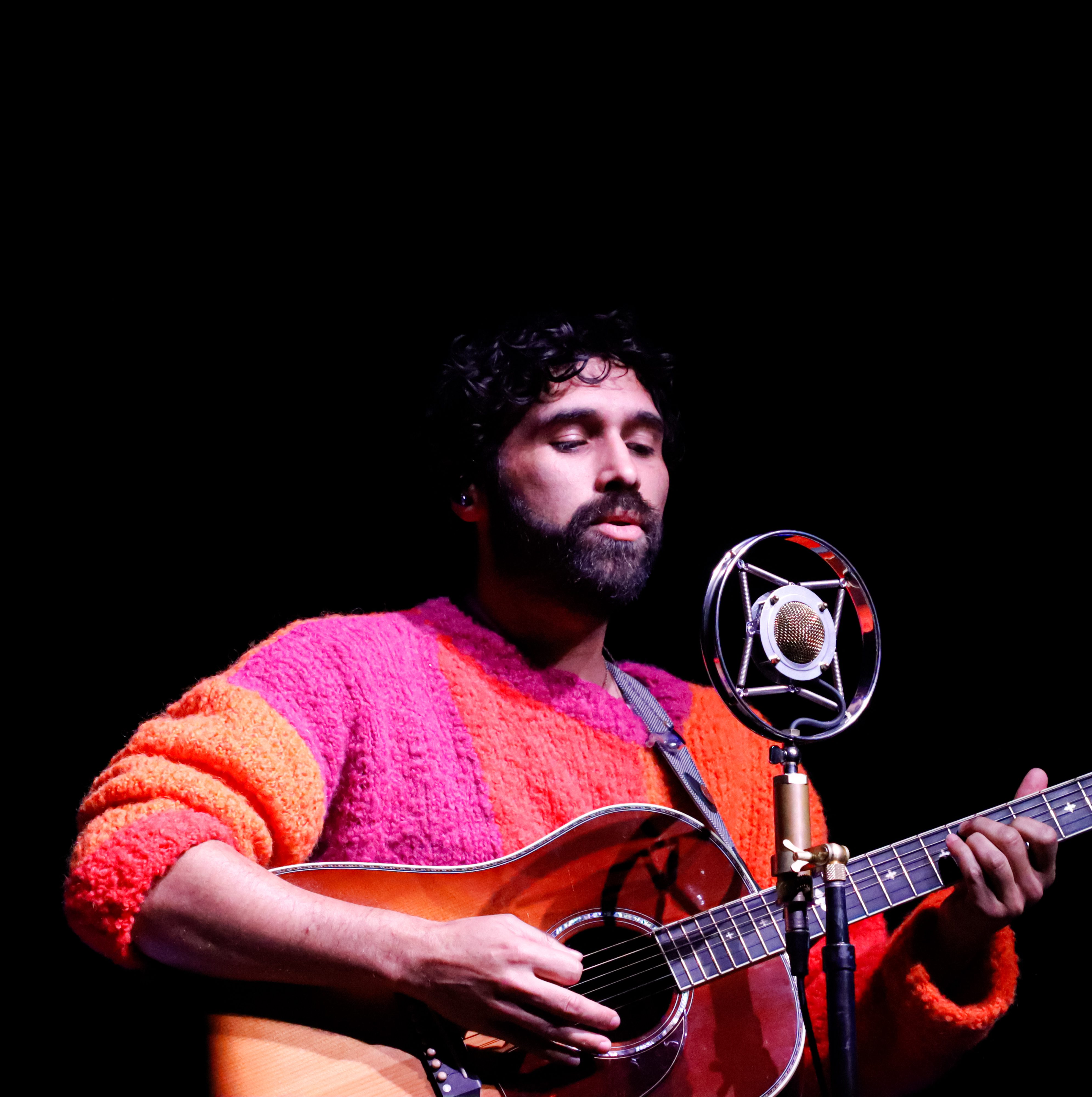 A musician with dark curly hair and a beard plays an acoustic guitar while singing into a vintage microphone, wearing a vibrant pink and orange sweater.