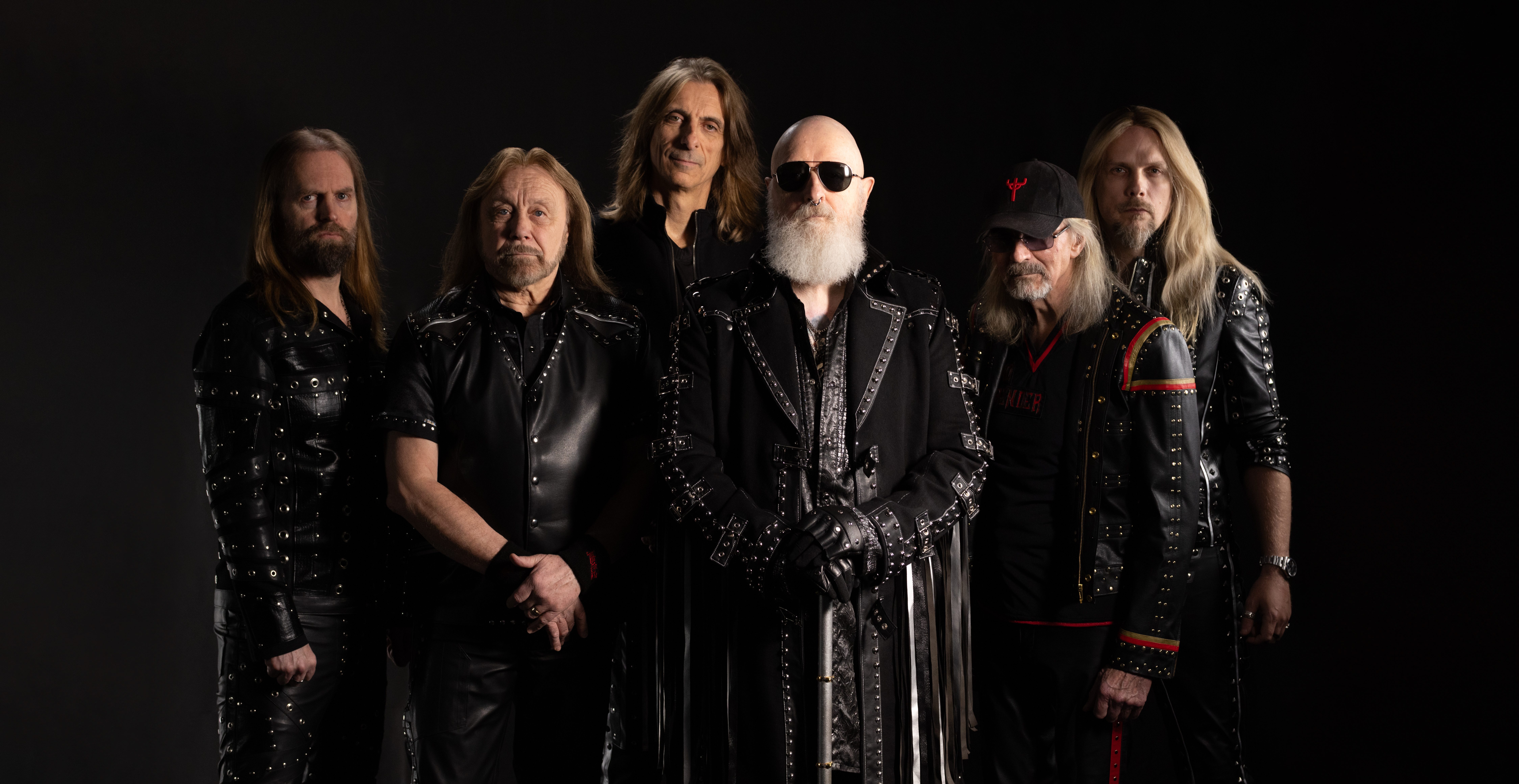 A group of six rock musicians posing against a dark background, wearing black leather outfits with metallic details.