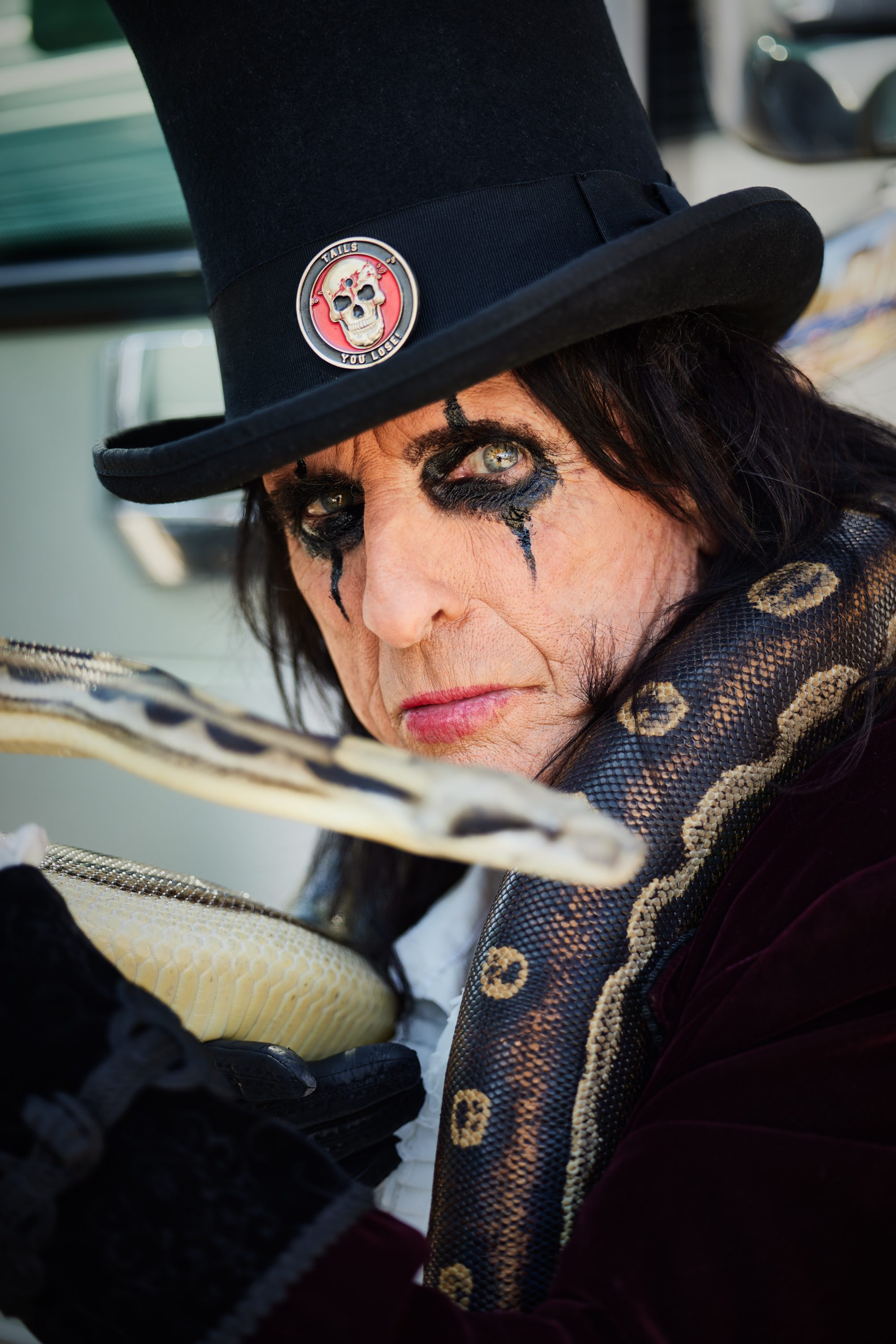 Close-up portrait of a man with dramatic makeup, wearing a black top hat and holding a snake, embodying a theatrical rock persona.