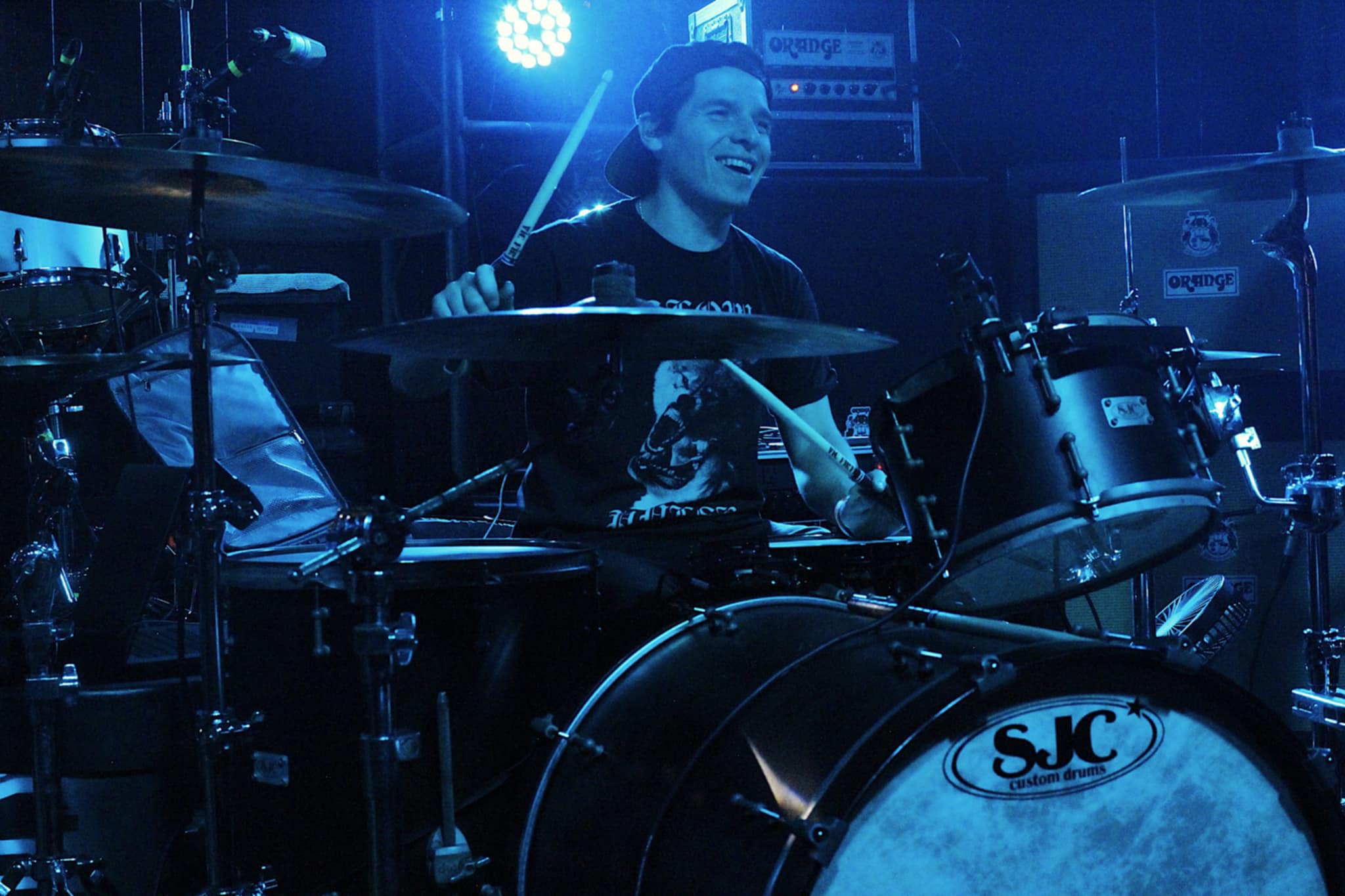 A drummer performs energetically on stage, surrounded by blue lighting, while playing a black drum set labeled with 'SJC Custom Drums'.
