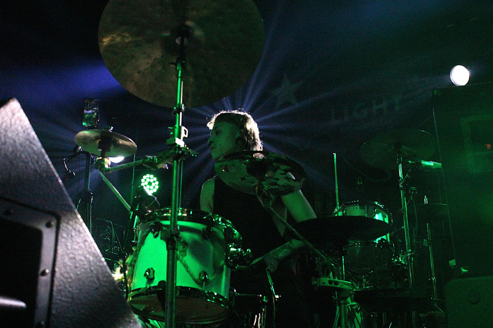 A drummer performing on stage with green lighting and dramatic backlighting during a live music concert.