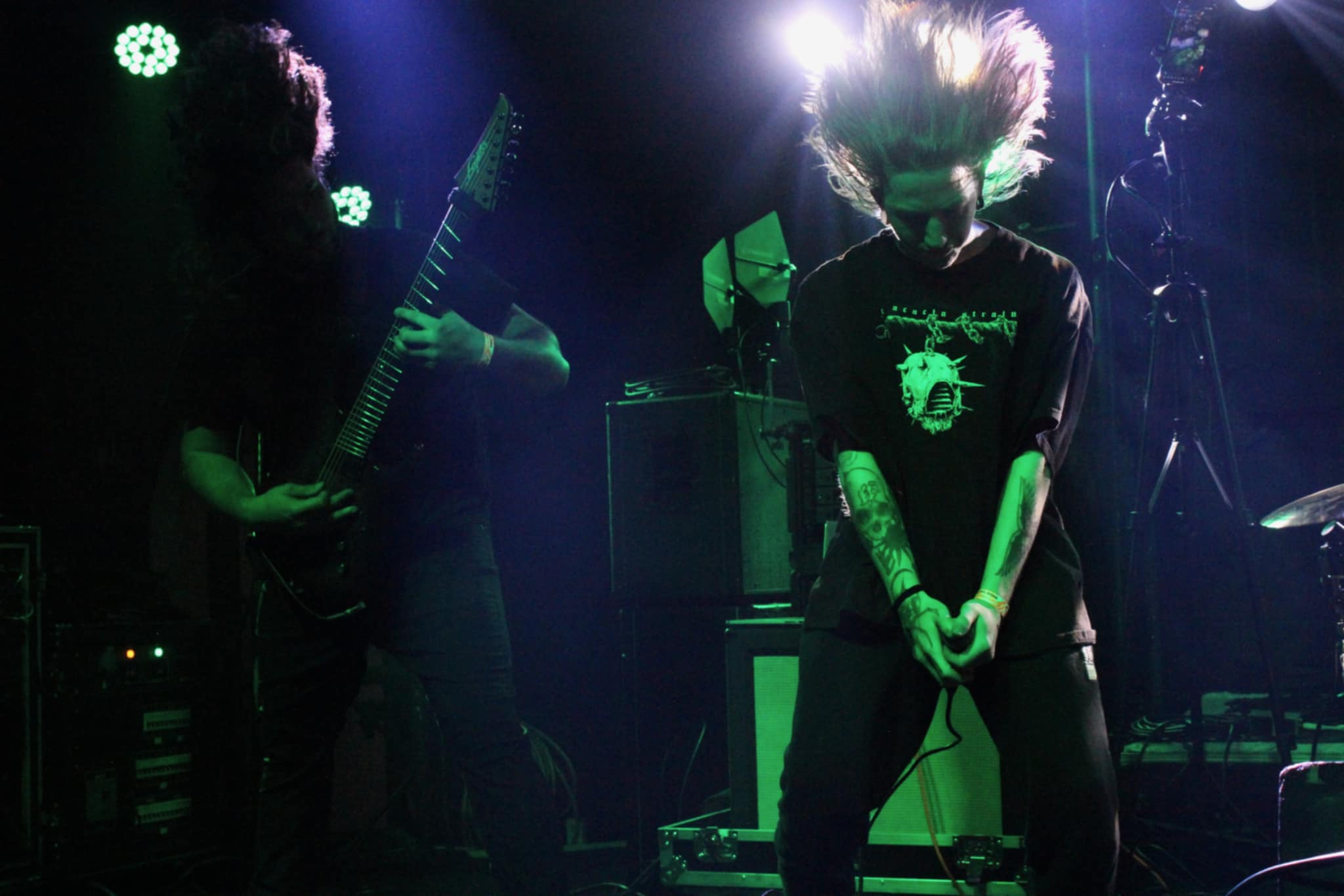 A live performance featuring two musicians in a dark, colorful setting. One is playing an electric guitar while the other is performing an intense posture, with their hair flying, suggesting vigorous movement and energy. The background is illuminated with stage lights, enhancing the lively atmosphere.