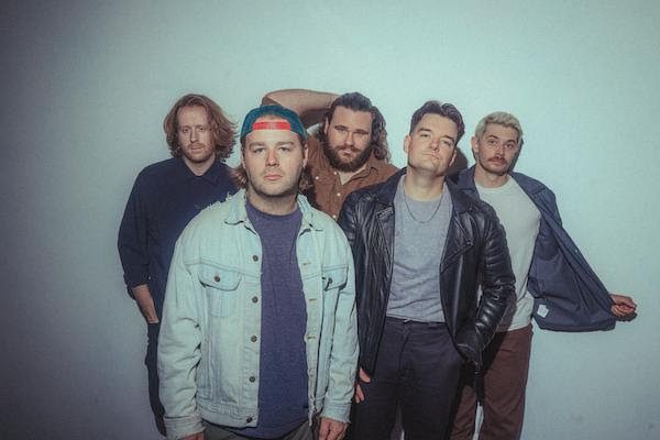 A five-member band posing for a photo, with one member in a denim jacket, another in a leather jacket, and others in casual attire, all against a plain background.