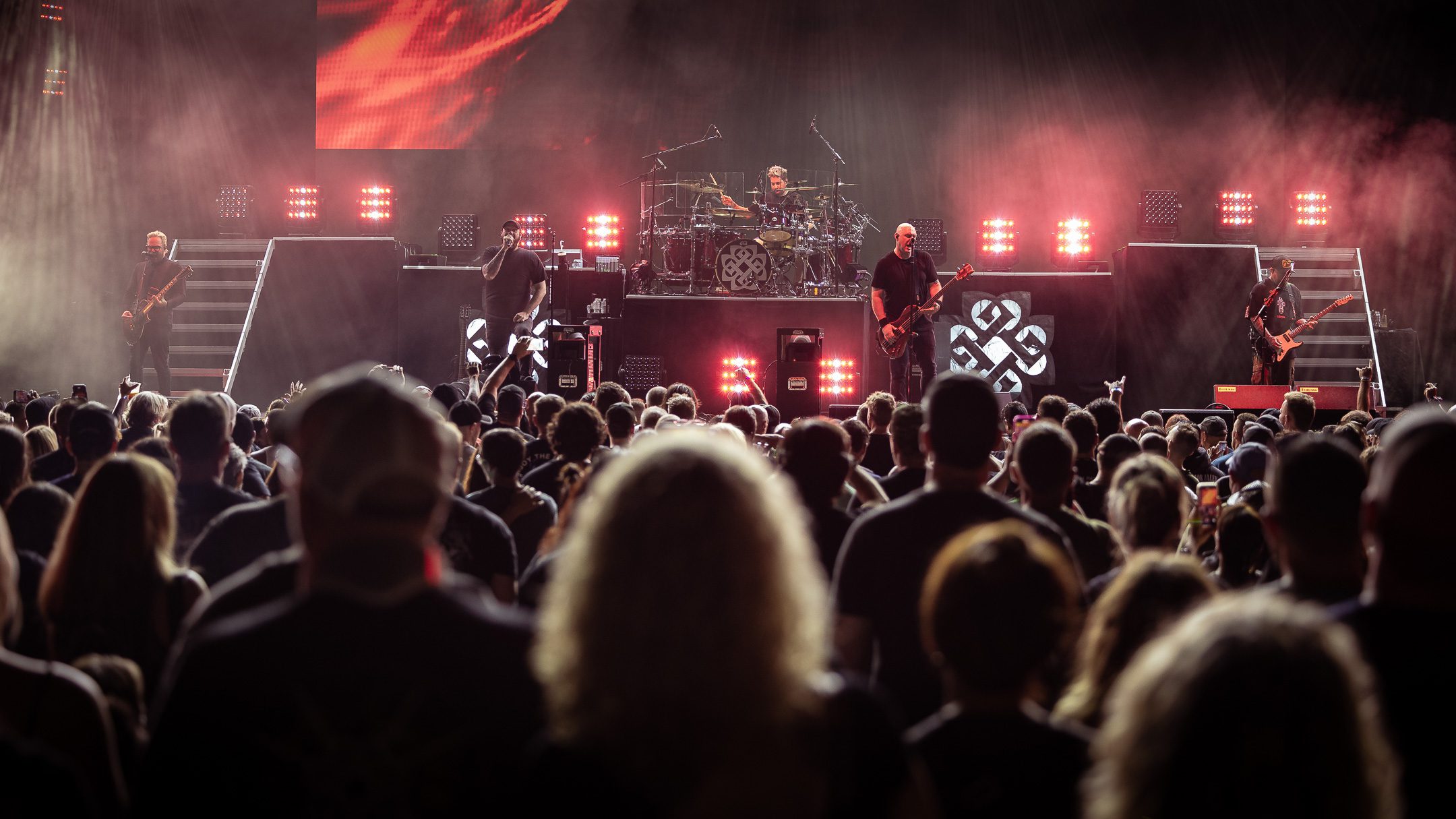 A live music performance featuring a band on stage with bright lights and a large audience in front.
