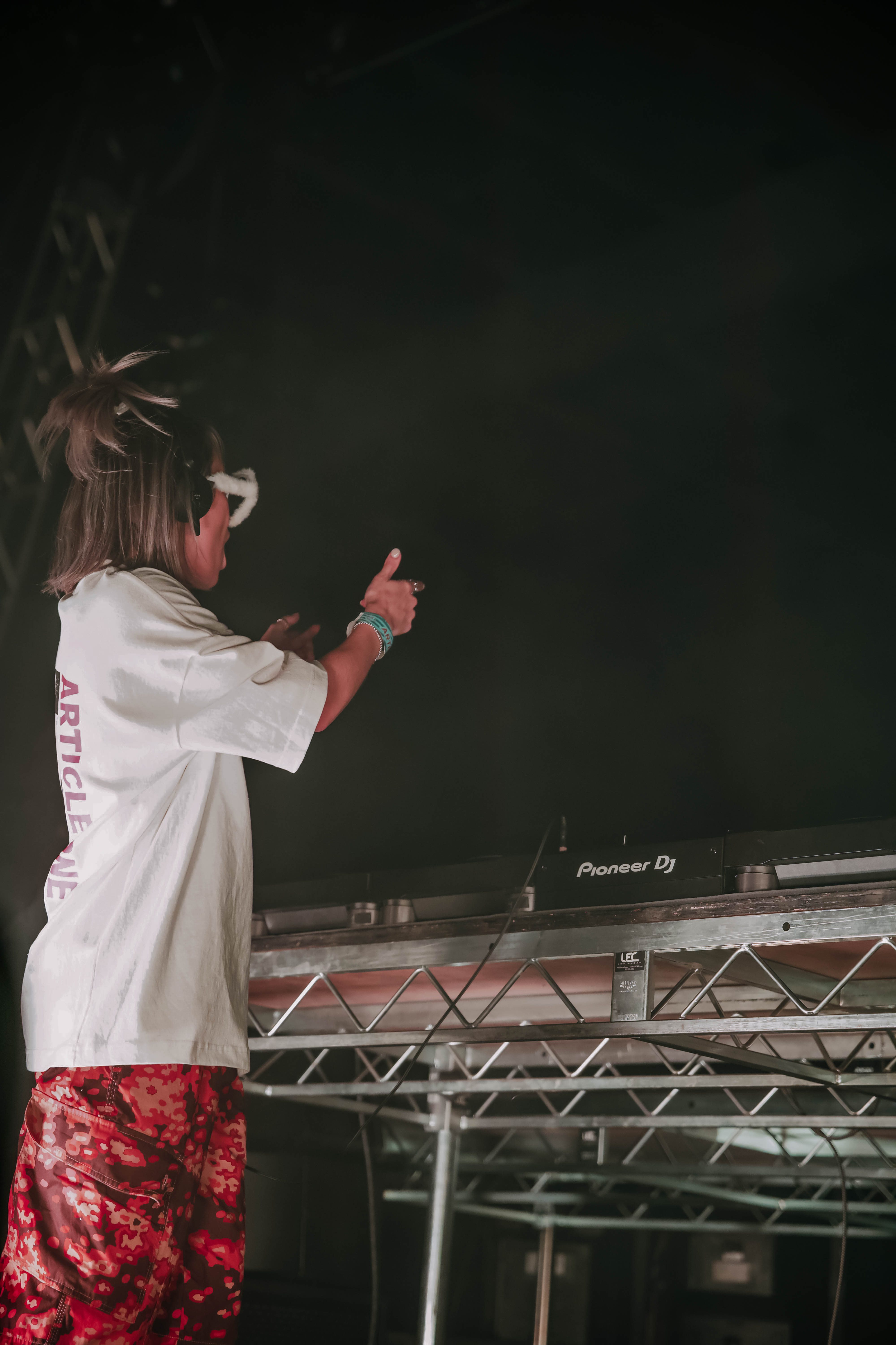 DJ performing on stage with a DJ mixer, wearing a white oversized shirt and red camouflage pants, showing enthusiasm during a set at a music festival.