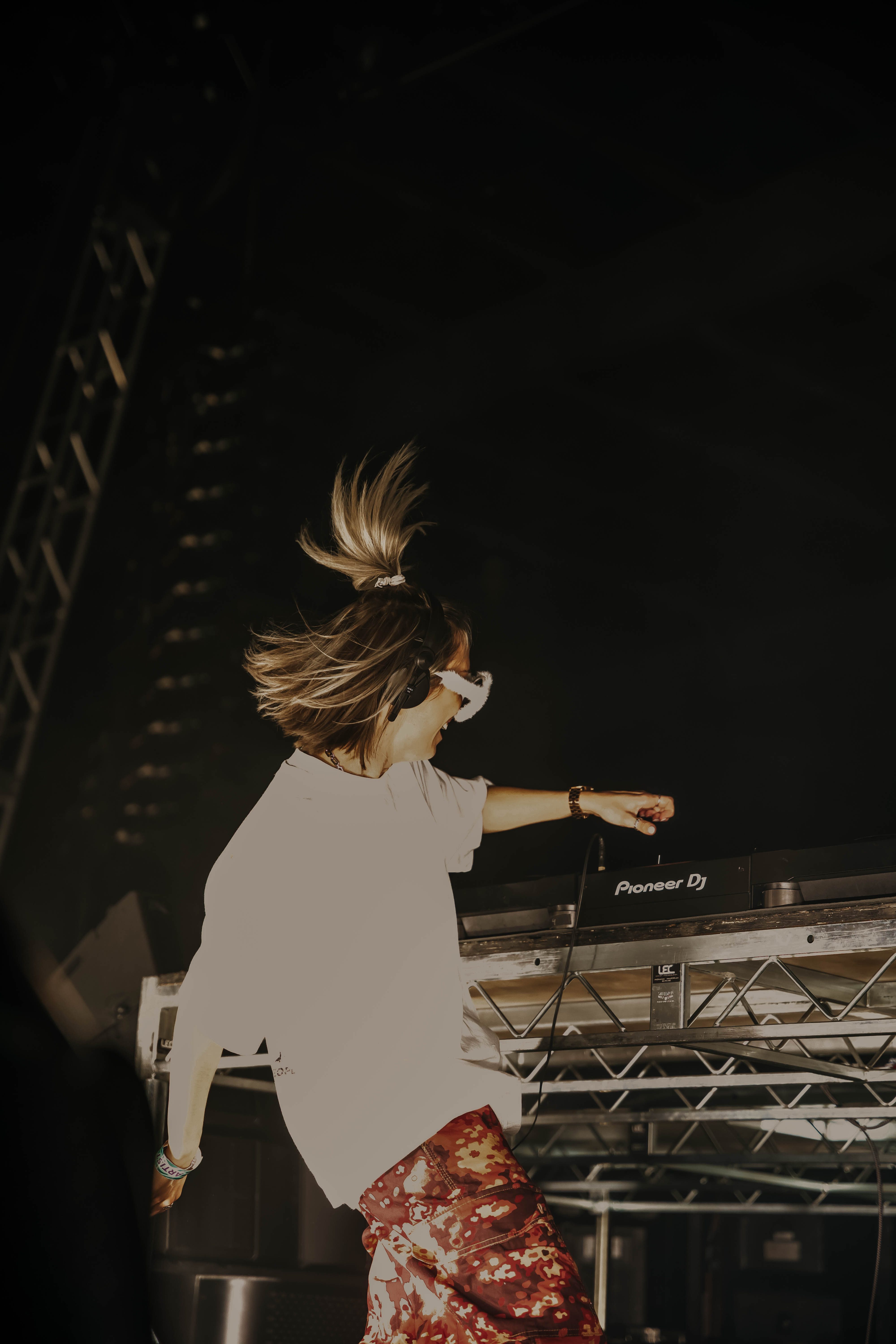 A DJ with blonde hair tied up is performing at a concert, engaging with the equipment on a stage. The stage is dimly lit, highlighting the DJ and the equipment.