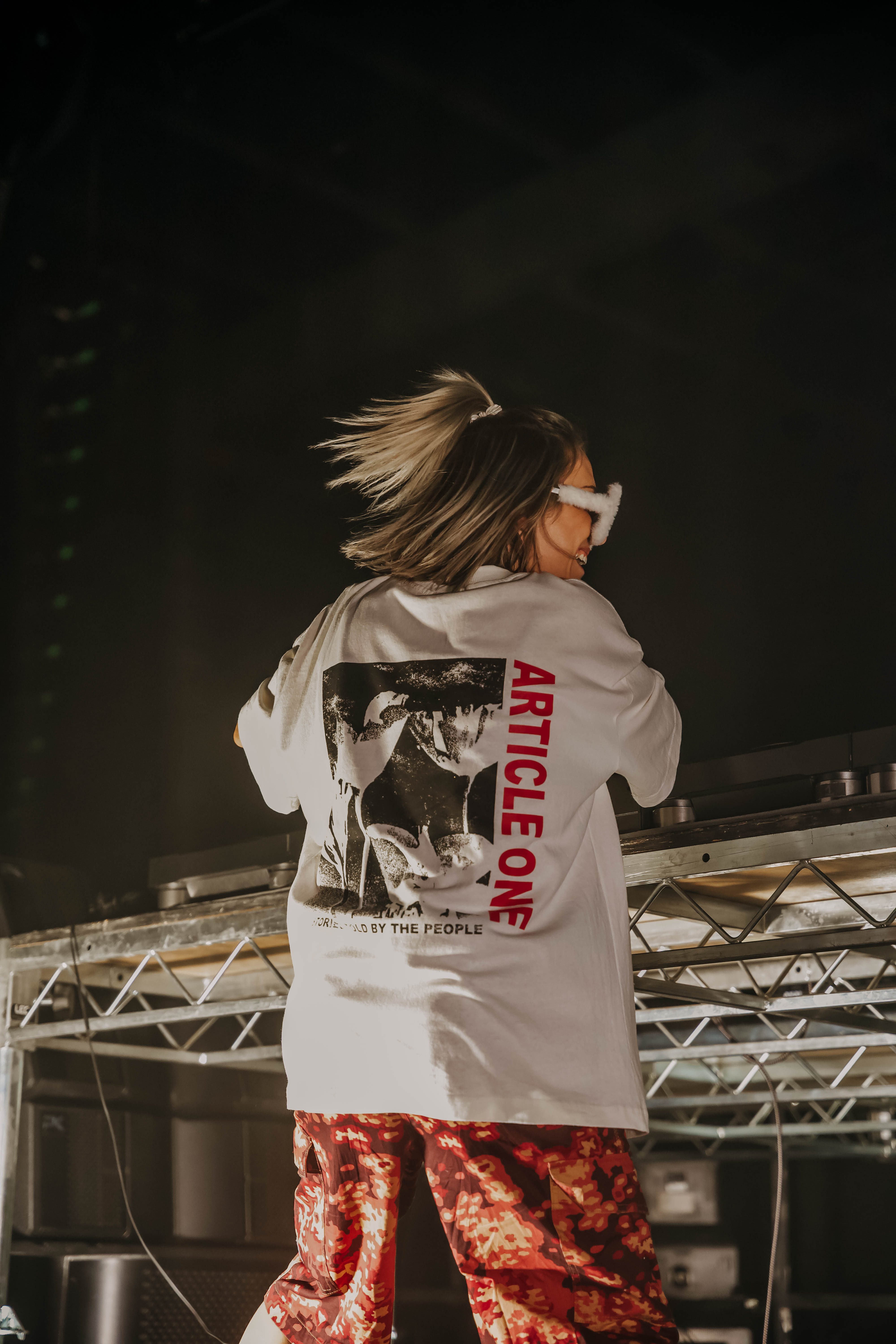 DJ Sippy performing at a music event, wearing a graphic t-shirt and red patterned shorts, with her back to the camera, showcasing her energetic performance.