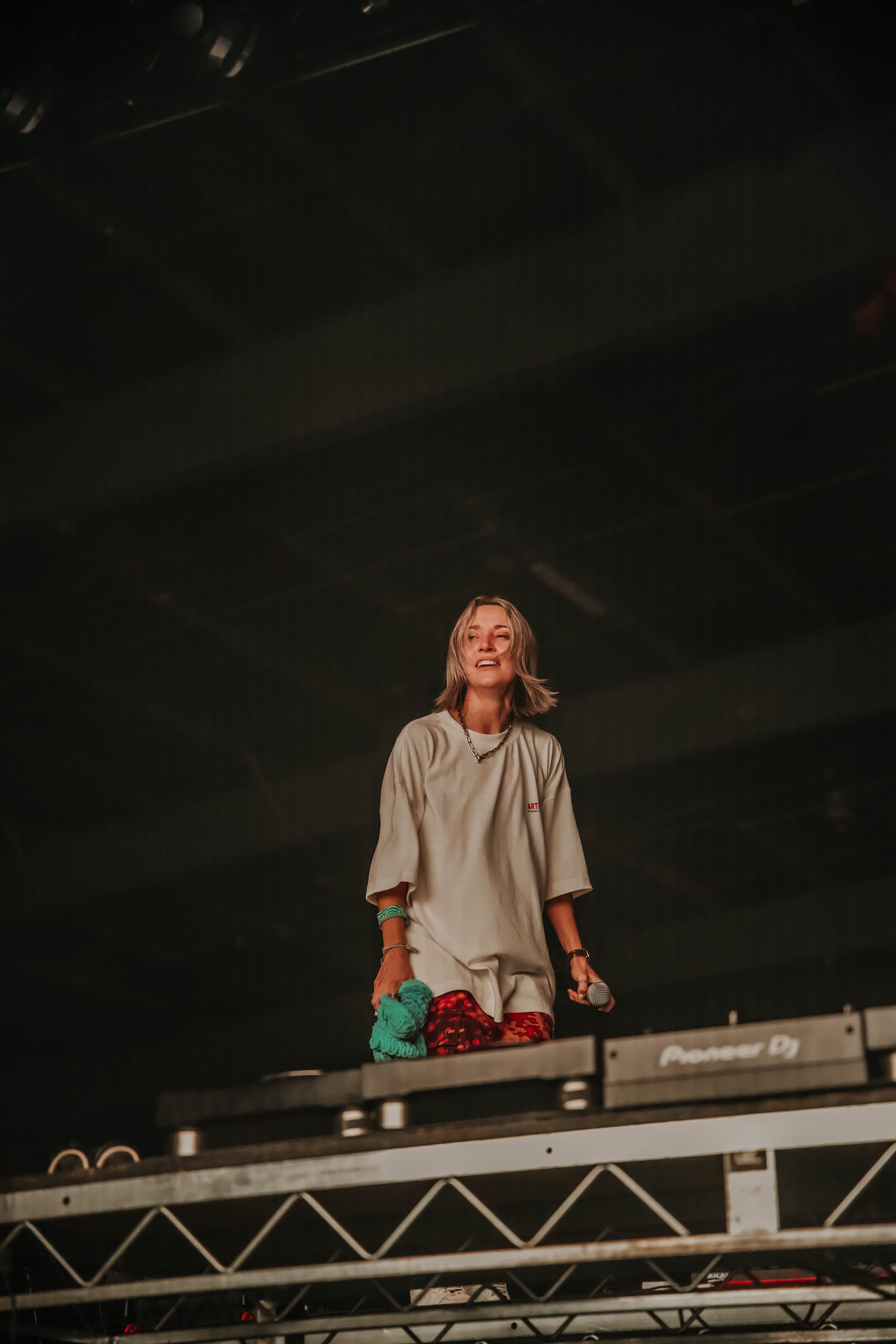 A female DJ performing on stage, smiling and engaging with the crowd while holding a microphone and a colorful cloth in her hand.