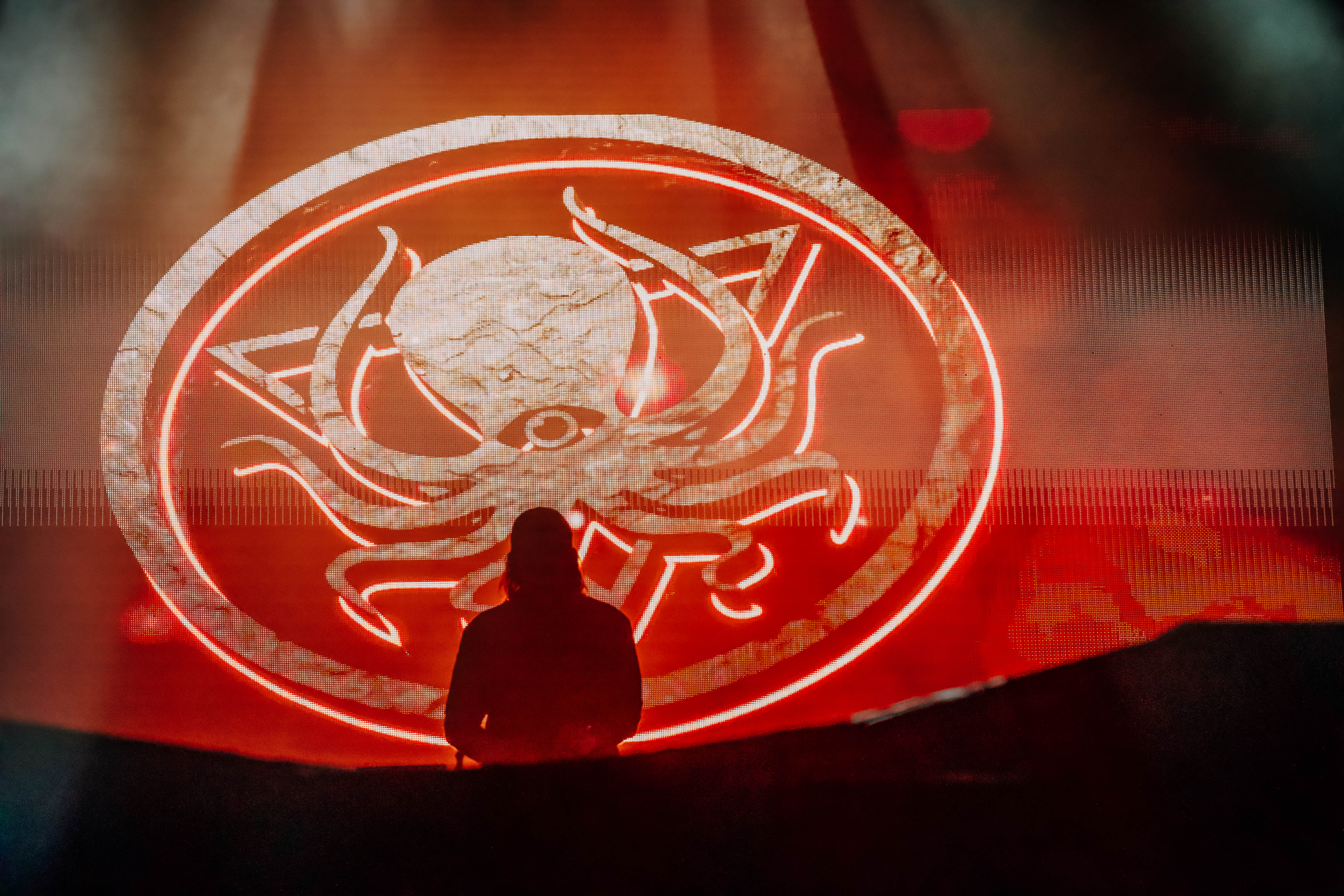 A DJ performs on stage silhouetted against a vibrant red backdrop featuring an octopus logo.