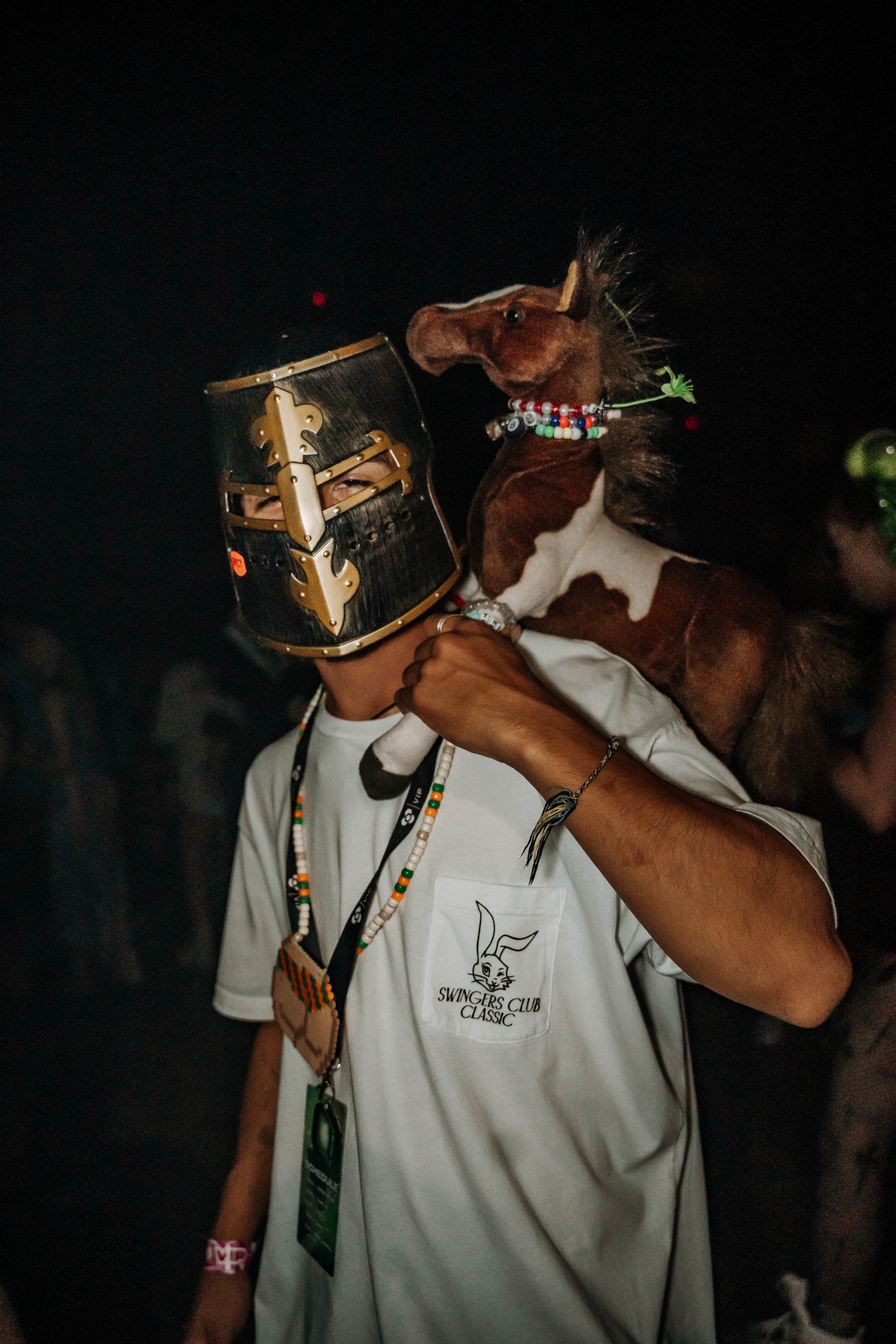 A person wearing a helmet and a white t-shirt holds a stuffed horse on their shoulder. They are surrounded by a dark party atmosphere, adorned with colorful accessories.
