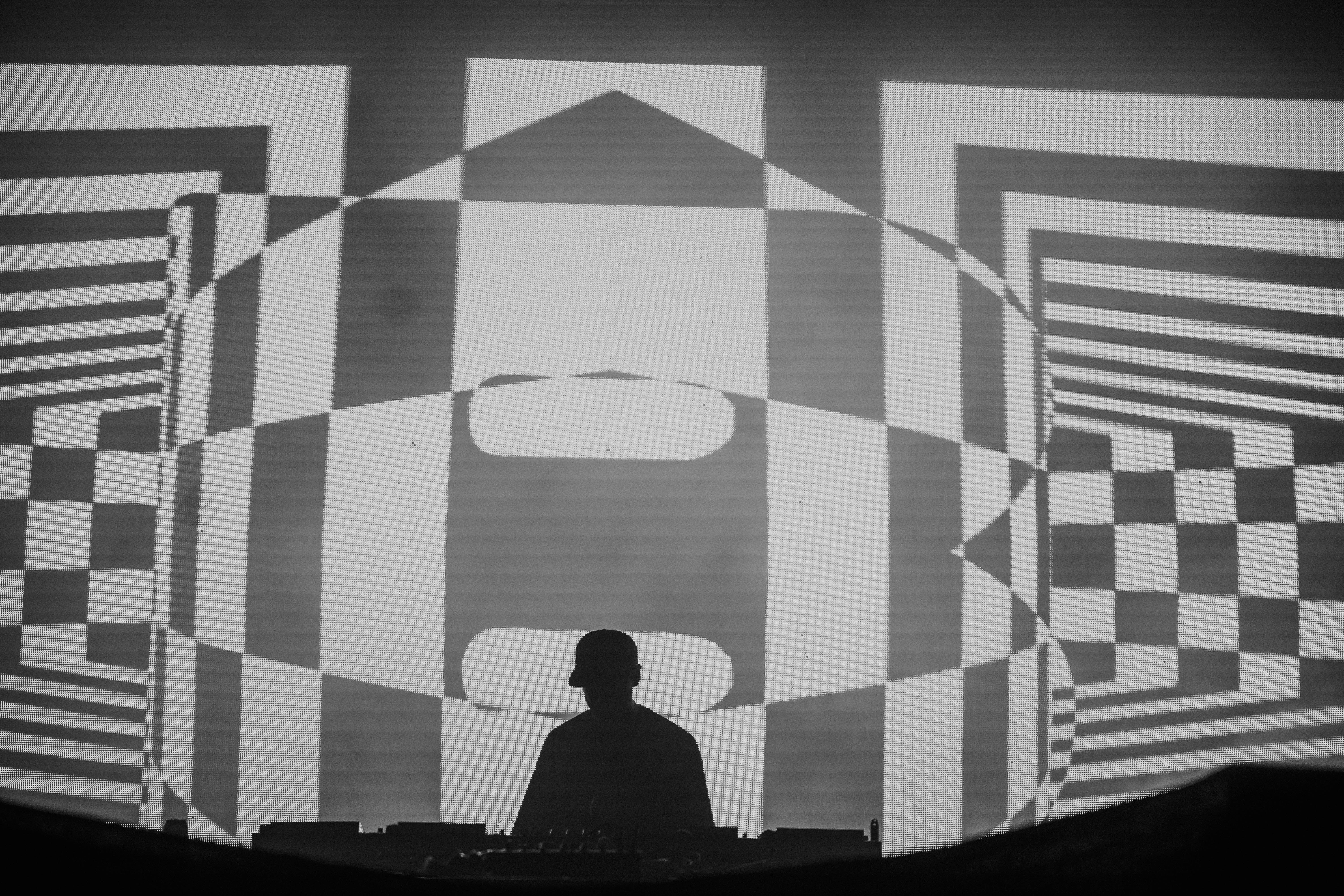 Silhouette of a DJ performing in front of psychedelic visuals on a large screen, featuring geometric patterns in black and white.