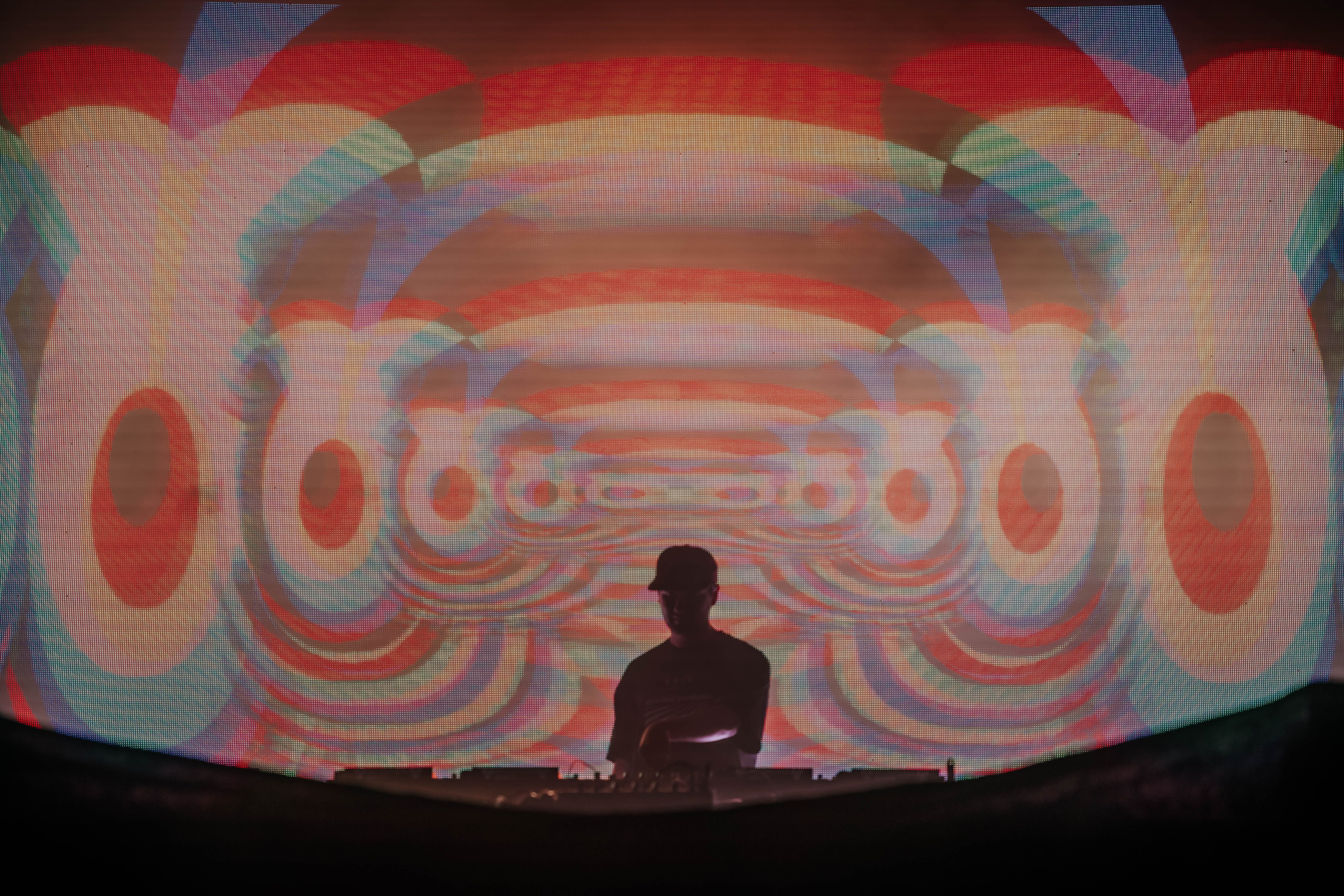 A DJ performs on stage with vibrant, colorful visuals projected behind, creating a mesmerizing atmosphere during a lively music event.