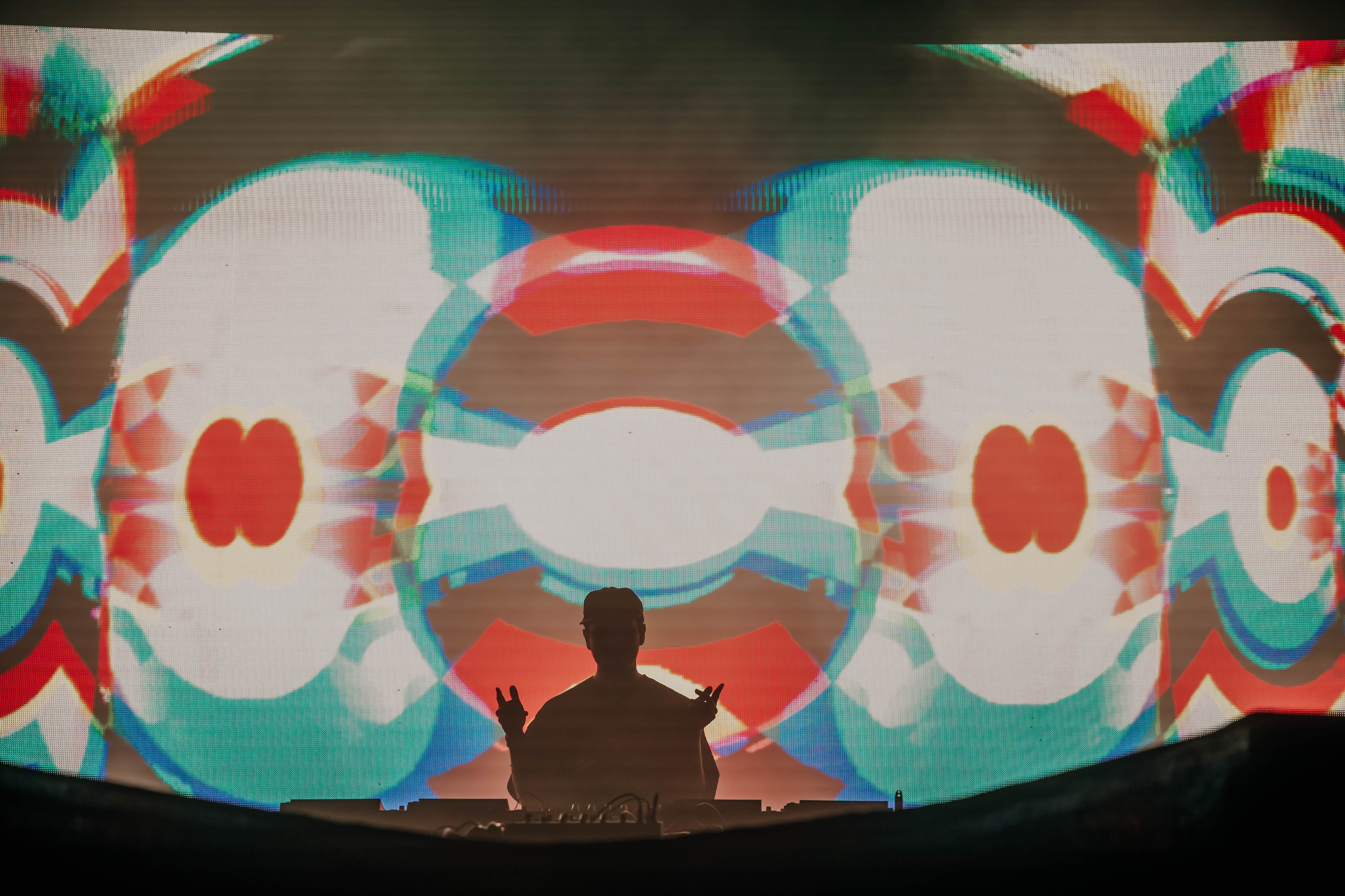 A DJ performing on stage, silhouetted against a vibrant, colorful visual display featuring abstract patterns.