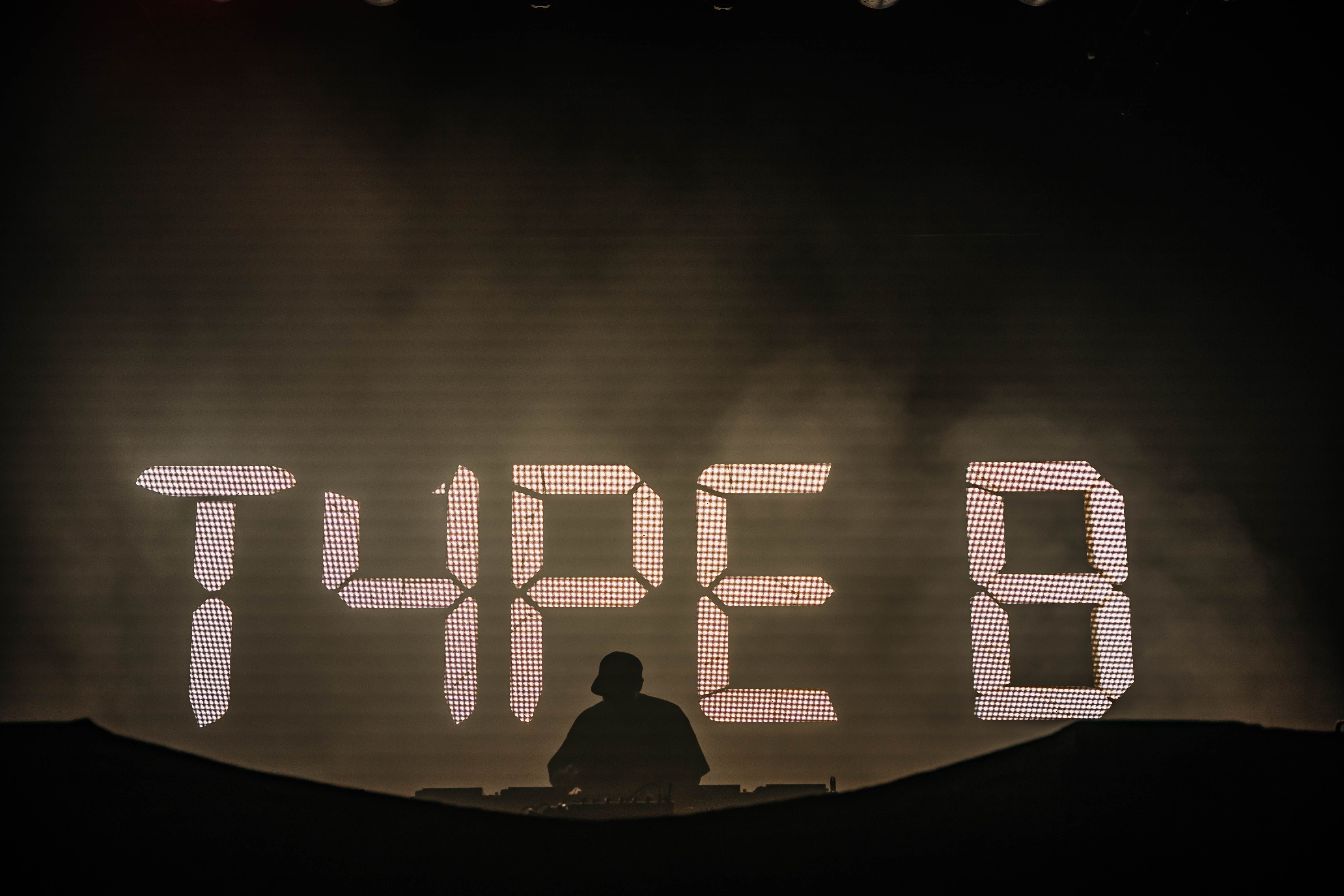 A DJ performing on stage, silhouetted against a large display that reads 'TYPE B' in illuminated numbers.