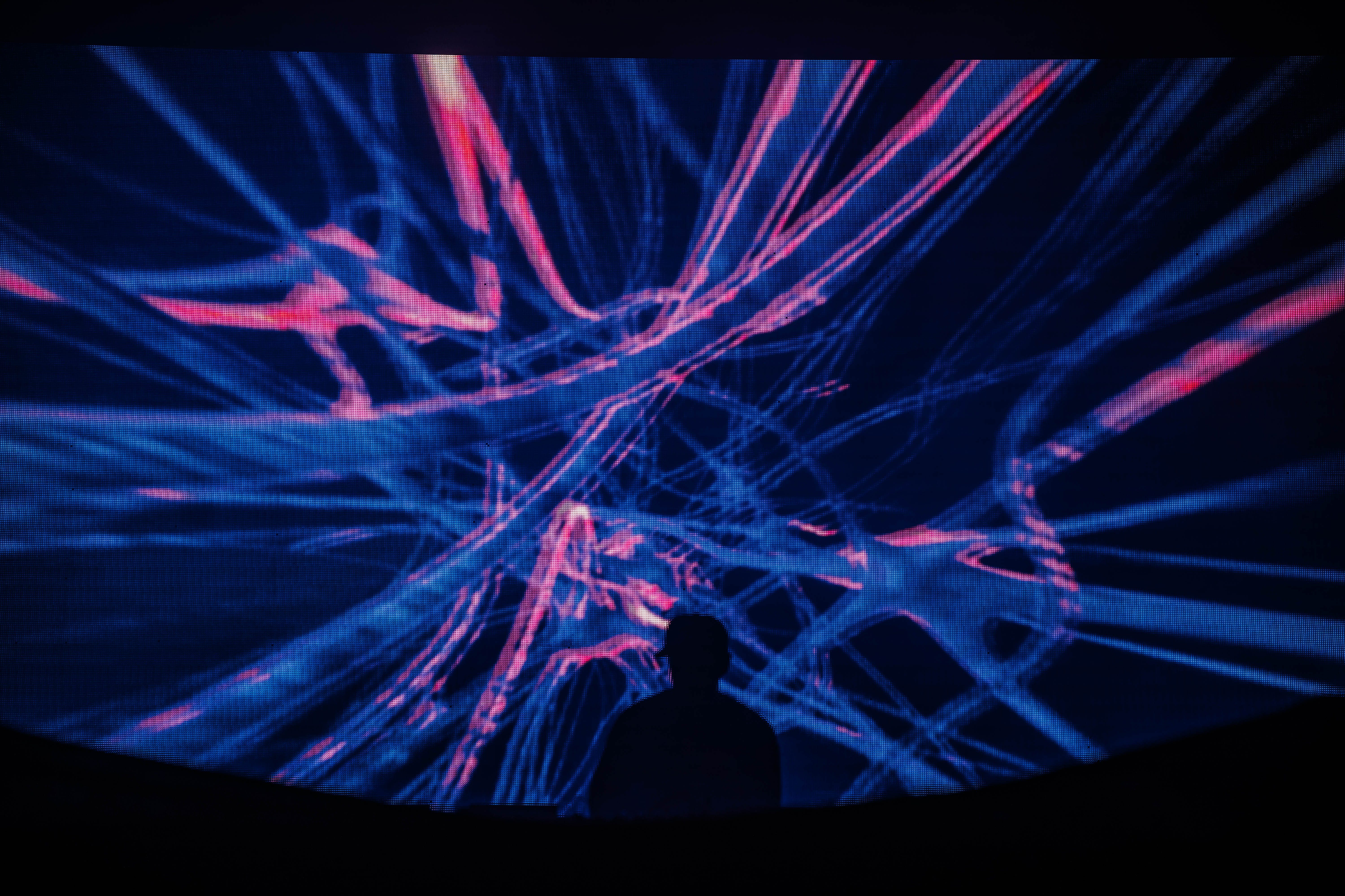 A silhouette of a DJ performing in front of a vibrant, abstract visual display with swirling blue and pink lines, creating a dynamic and energetic atmosphere.