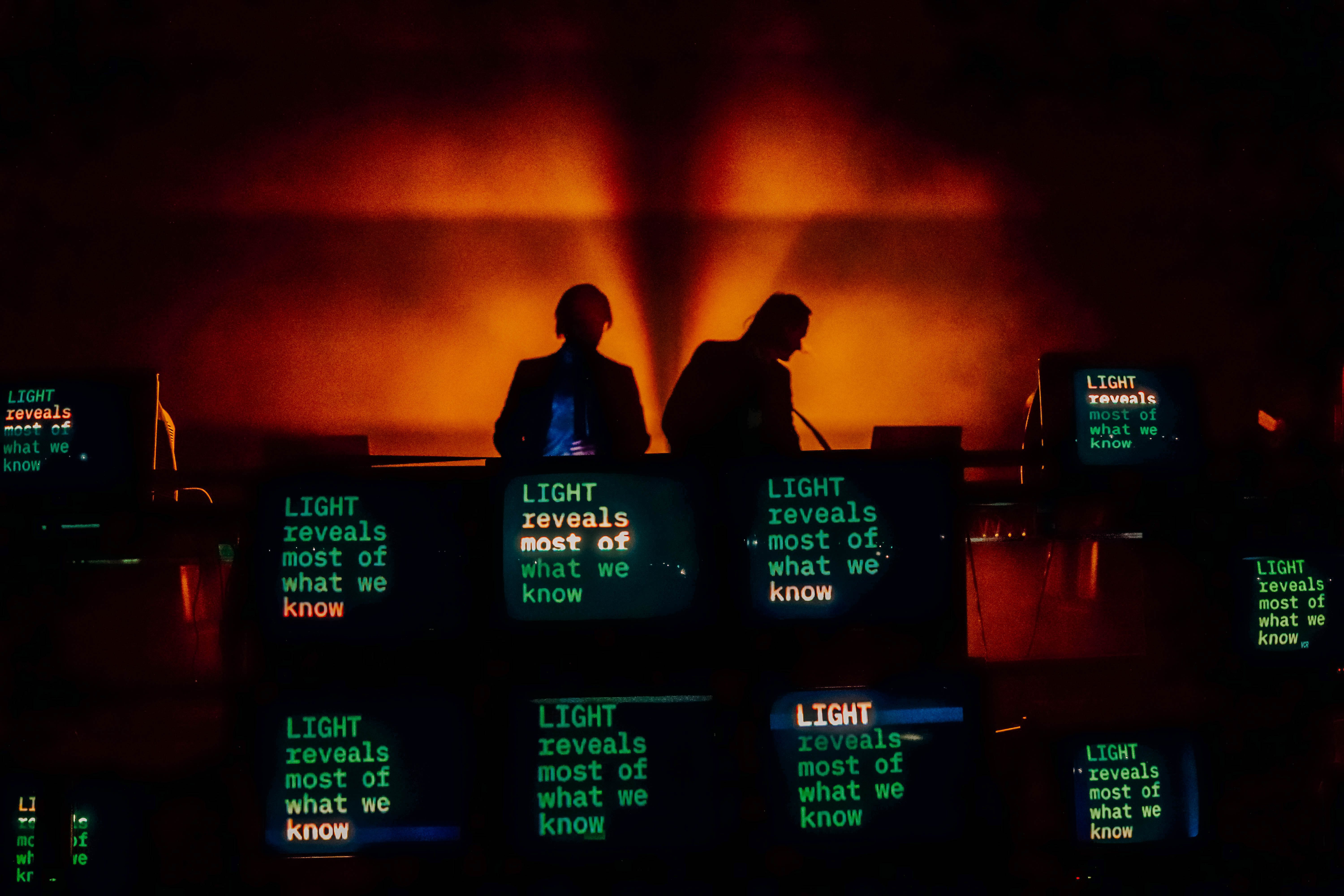 A dimly lit stage featuring two silhouetted DJs behind a lineup of vintage televisions displaying the text 'LIGHT reveals most of what we know' against an orange backdrop.