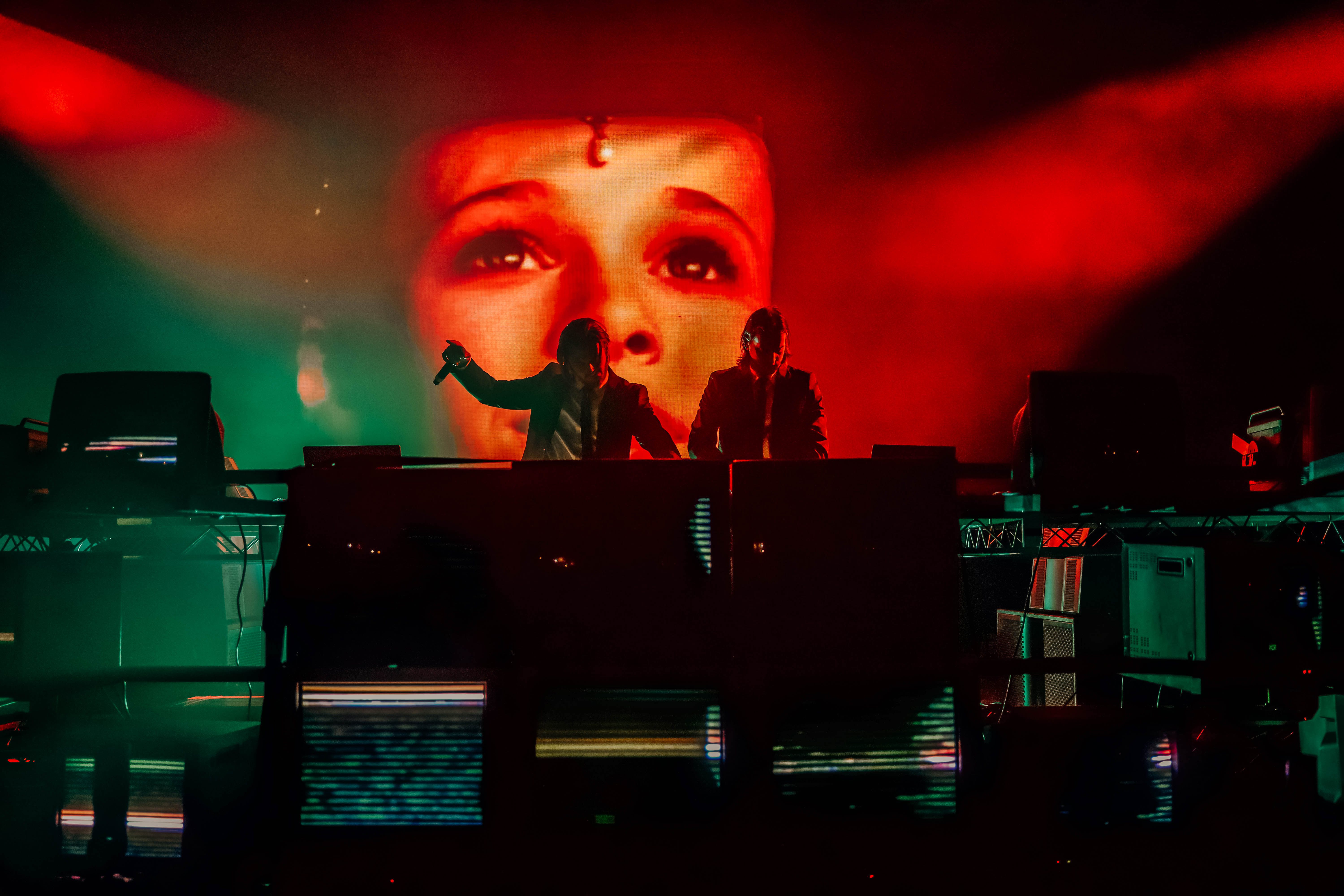 A vibrant concert scene featuring two DJs performing on stage with illuminated vintage TVs in front, set against a backdrop of colorful lights and a large projected image of a face.