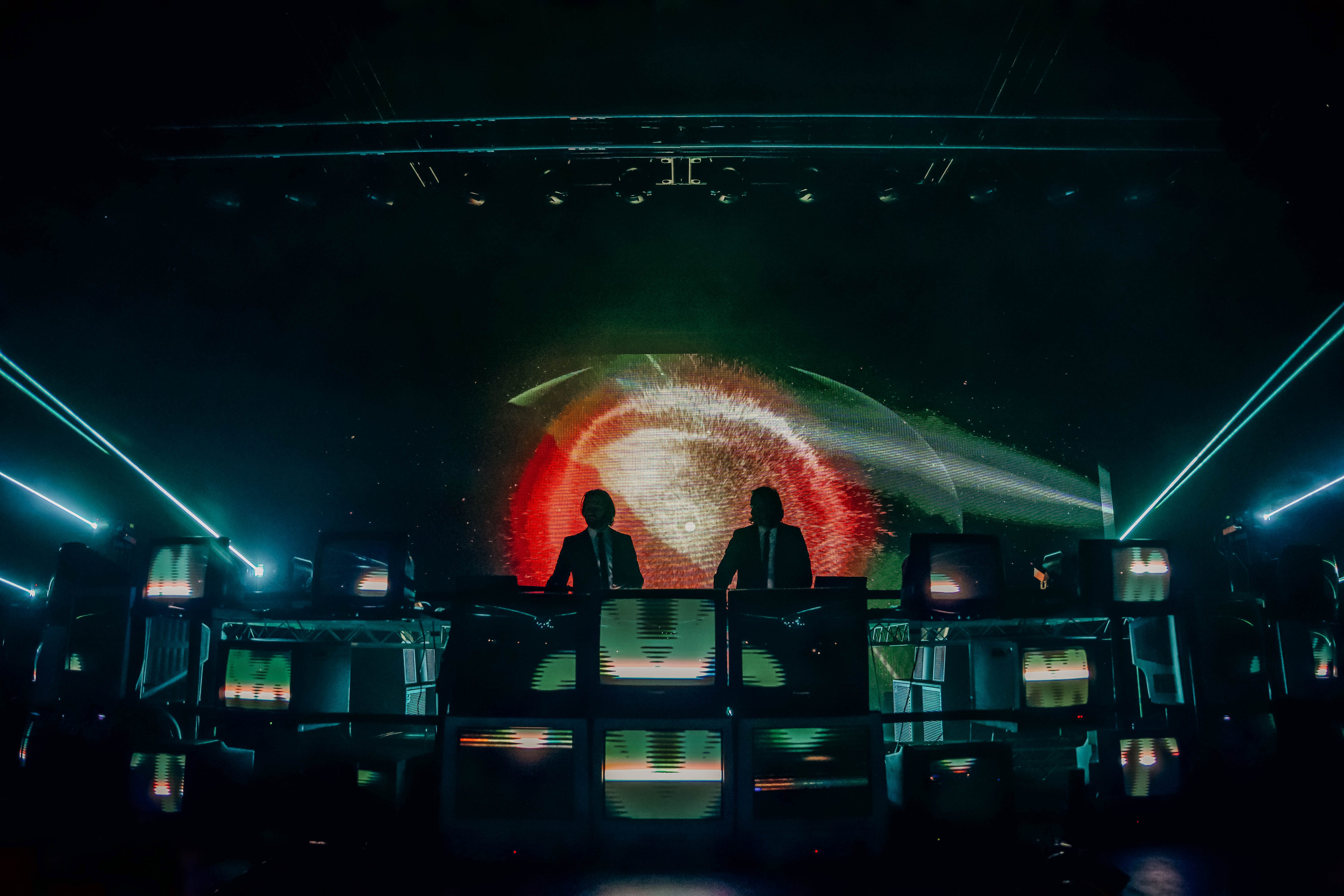 A DJ duo performing on stage surrounded by vintage televisions, with vibrant visuals displayed on a large screen behind them.
