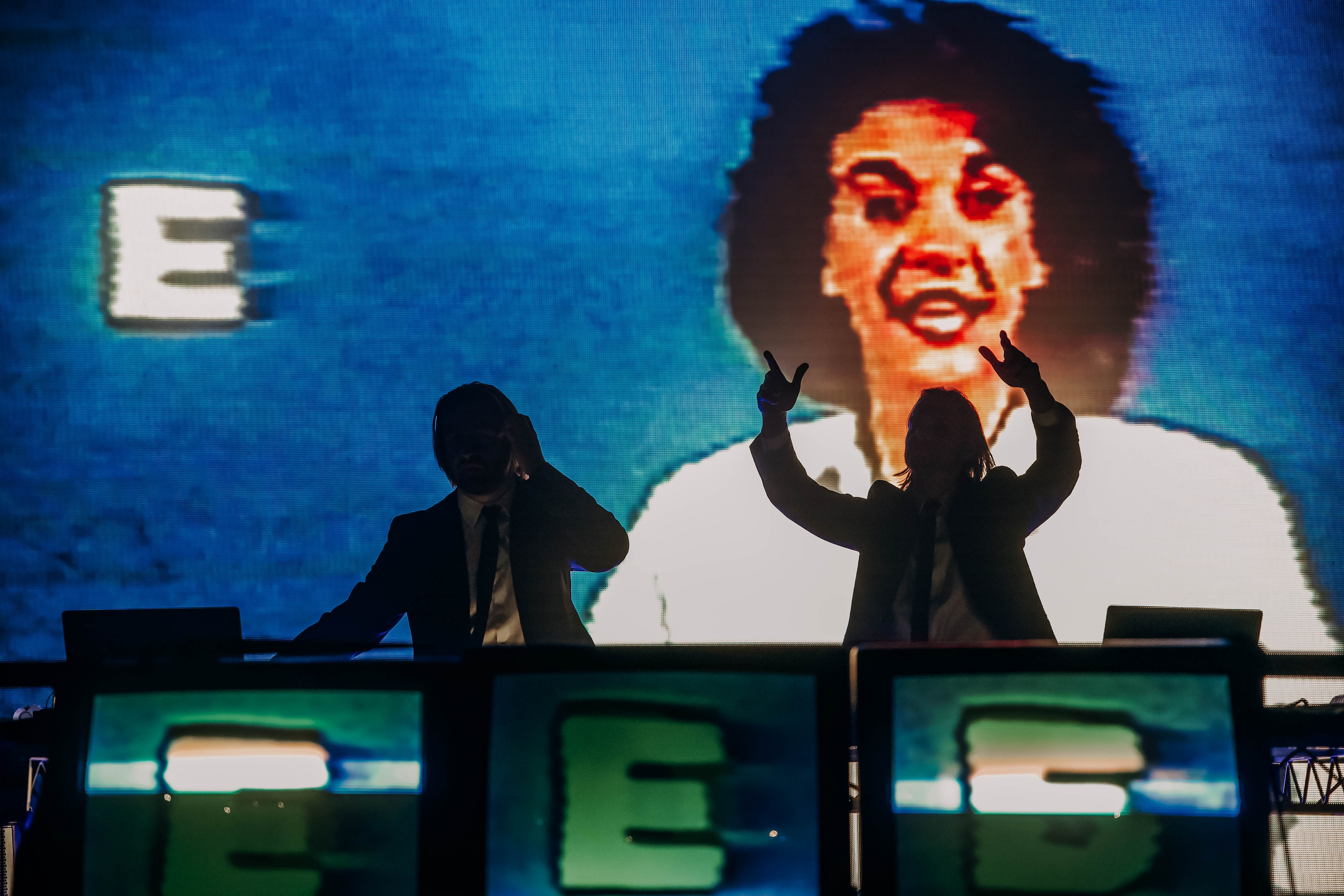 Two silhouetted DJs performing at a concert, with vibrant visuals projected in the background featuring a retro-style image.