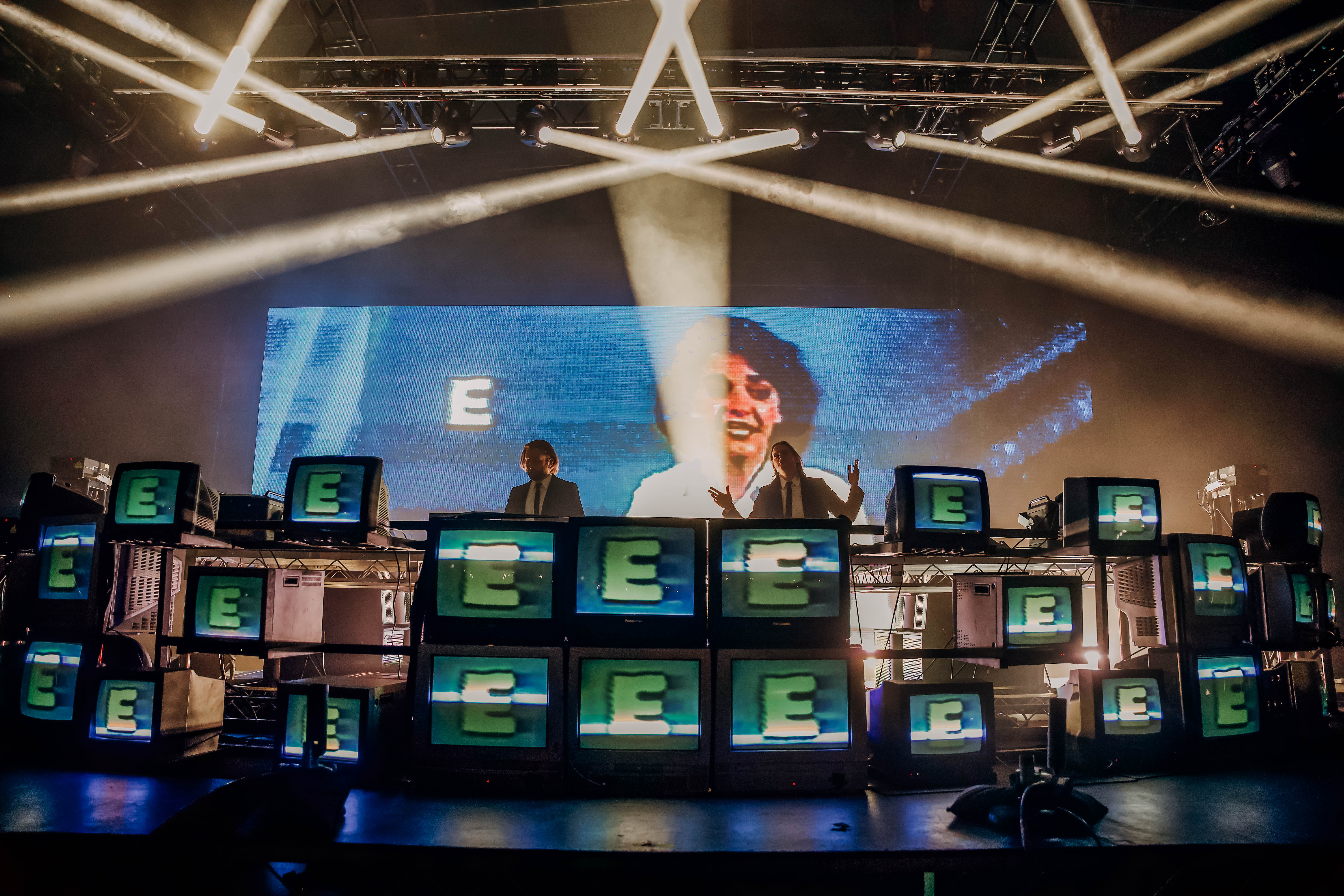 DJ duo performing live on stage with a backdrop of vintage TVs displaying visuals and beams of light shining down.