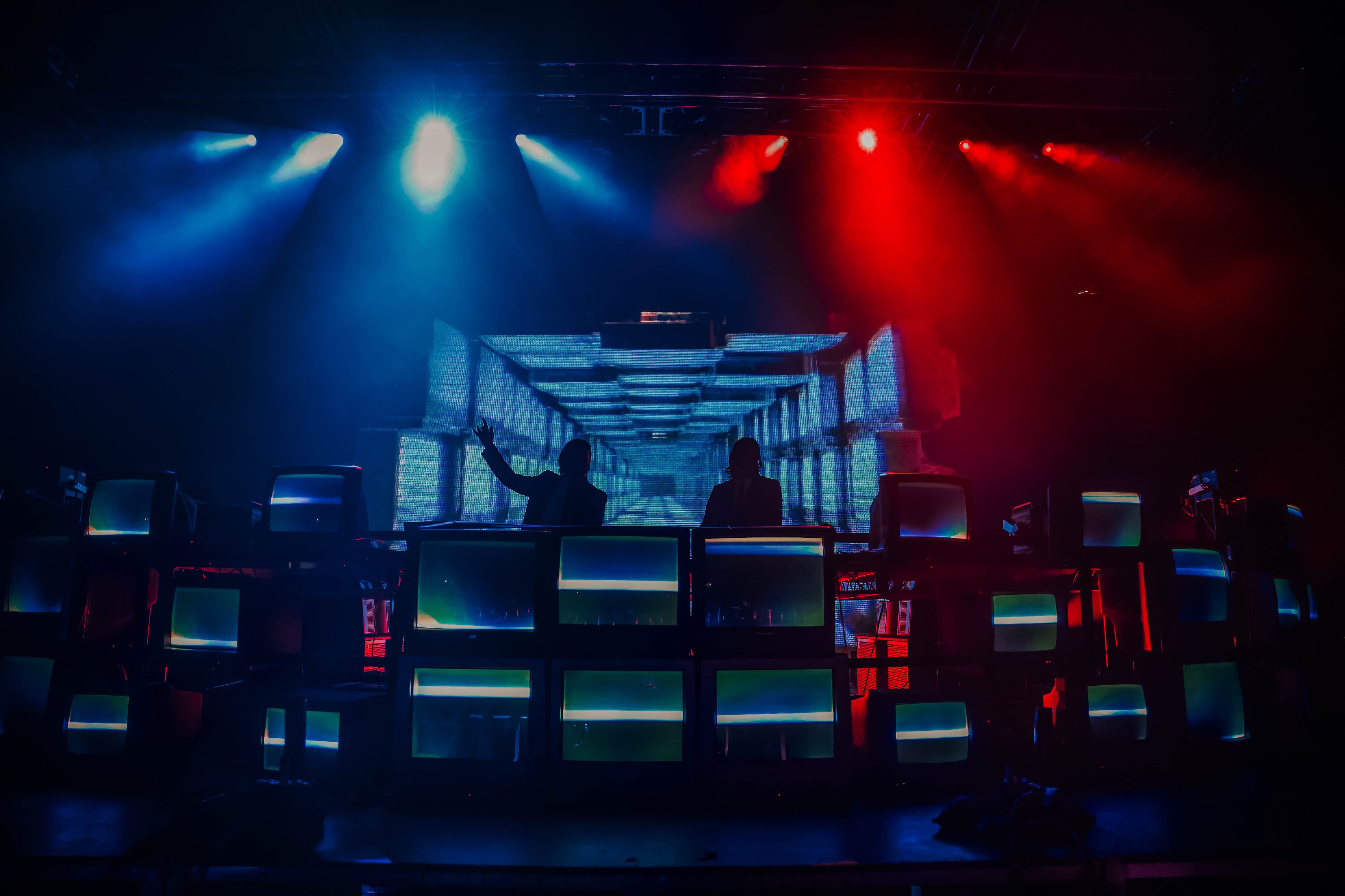 A DJ duo performing on stage with a backdrop of vintage TVs displaying colorful visuals under dynamic blue and red stage lights.