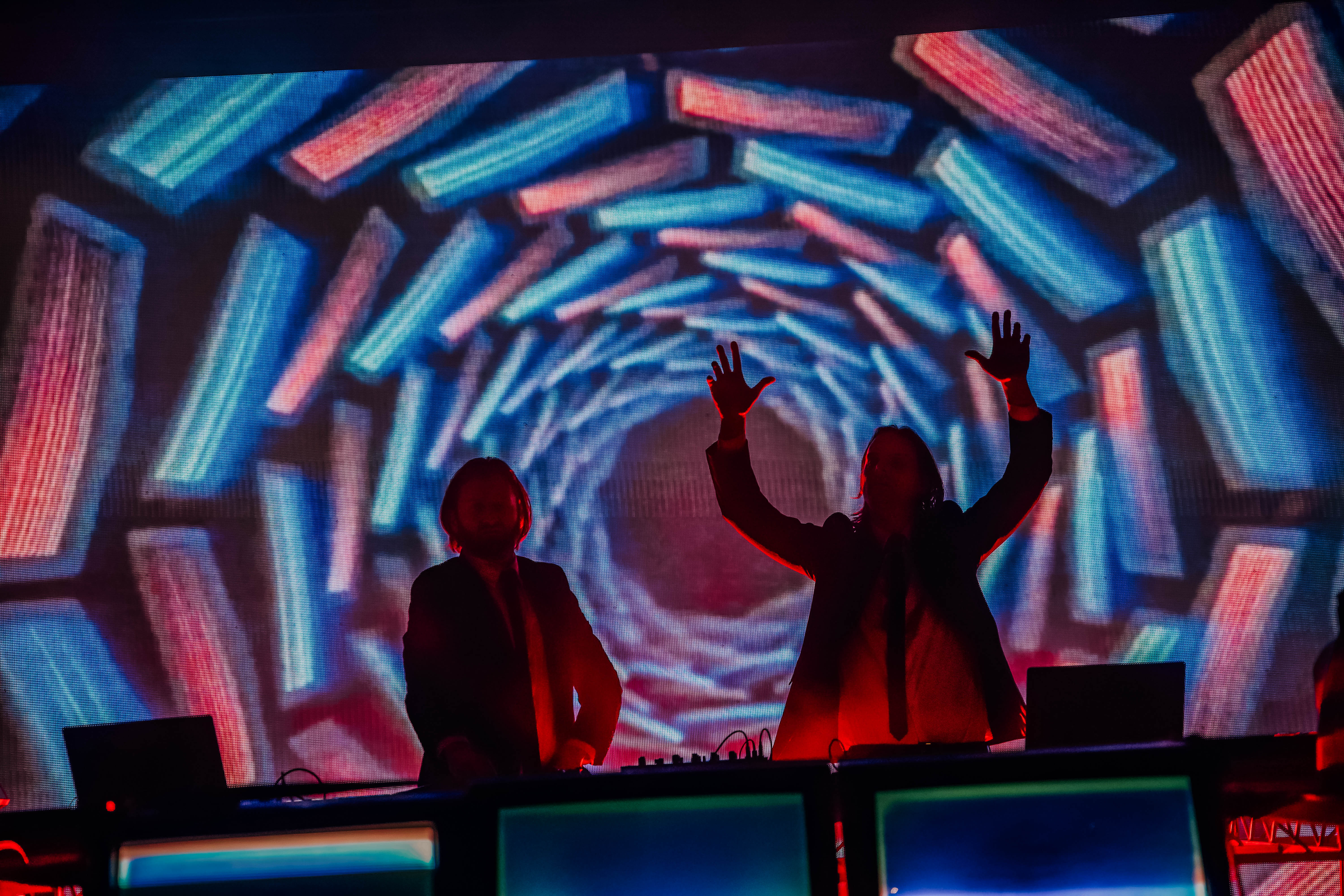 Two DJs performing on stage with hands raised, illuminated by vibrant, colorful visuals on a large screen behind them.