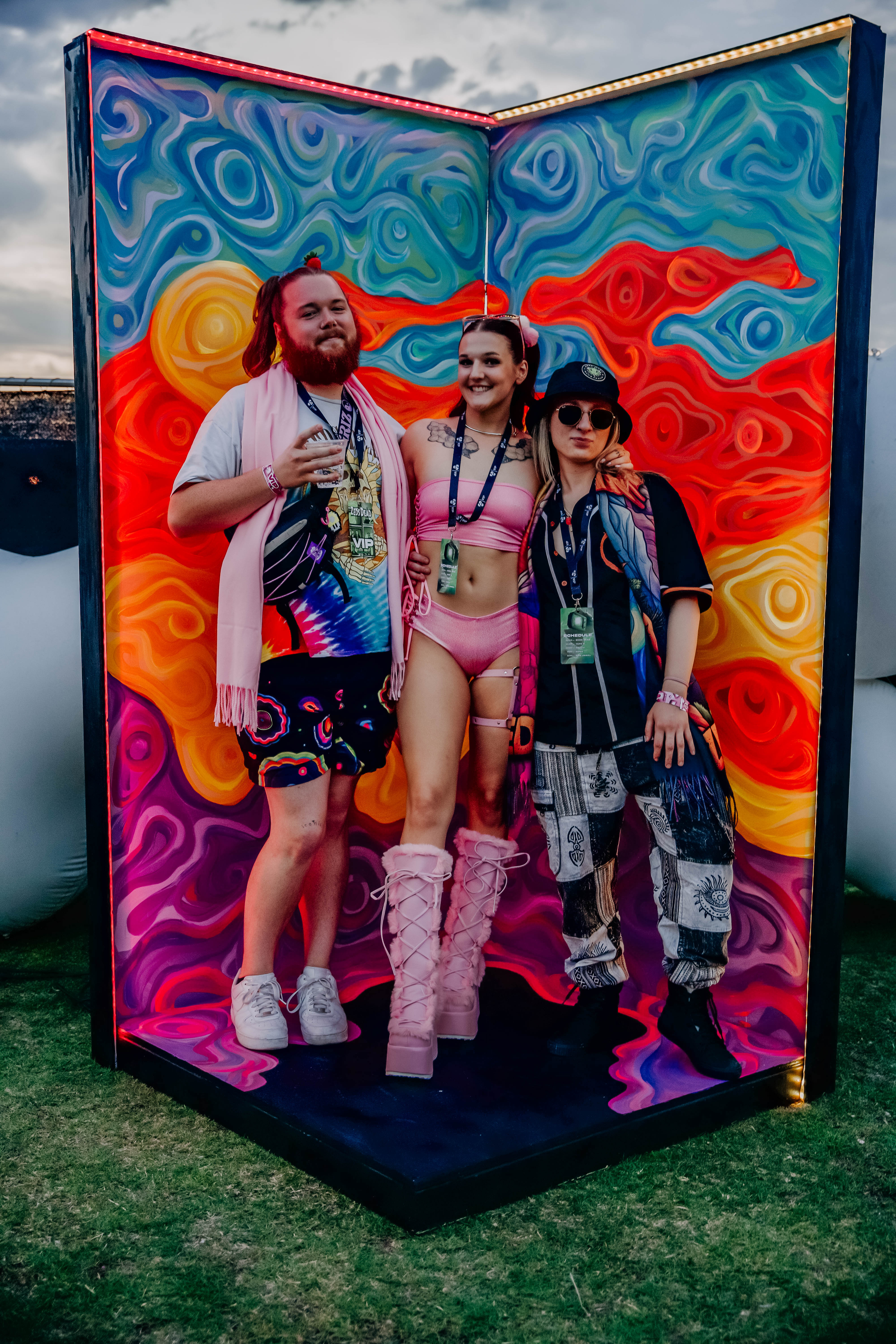 Three festival attendees pose in front of a colorful, swirling backdrop, showcasing vibrant psychedelic patterns. They display a mix of unique fashion styles and accessories, capturing the lively spirit of the event.