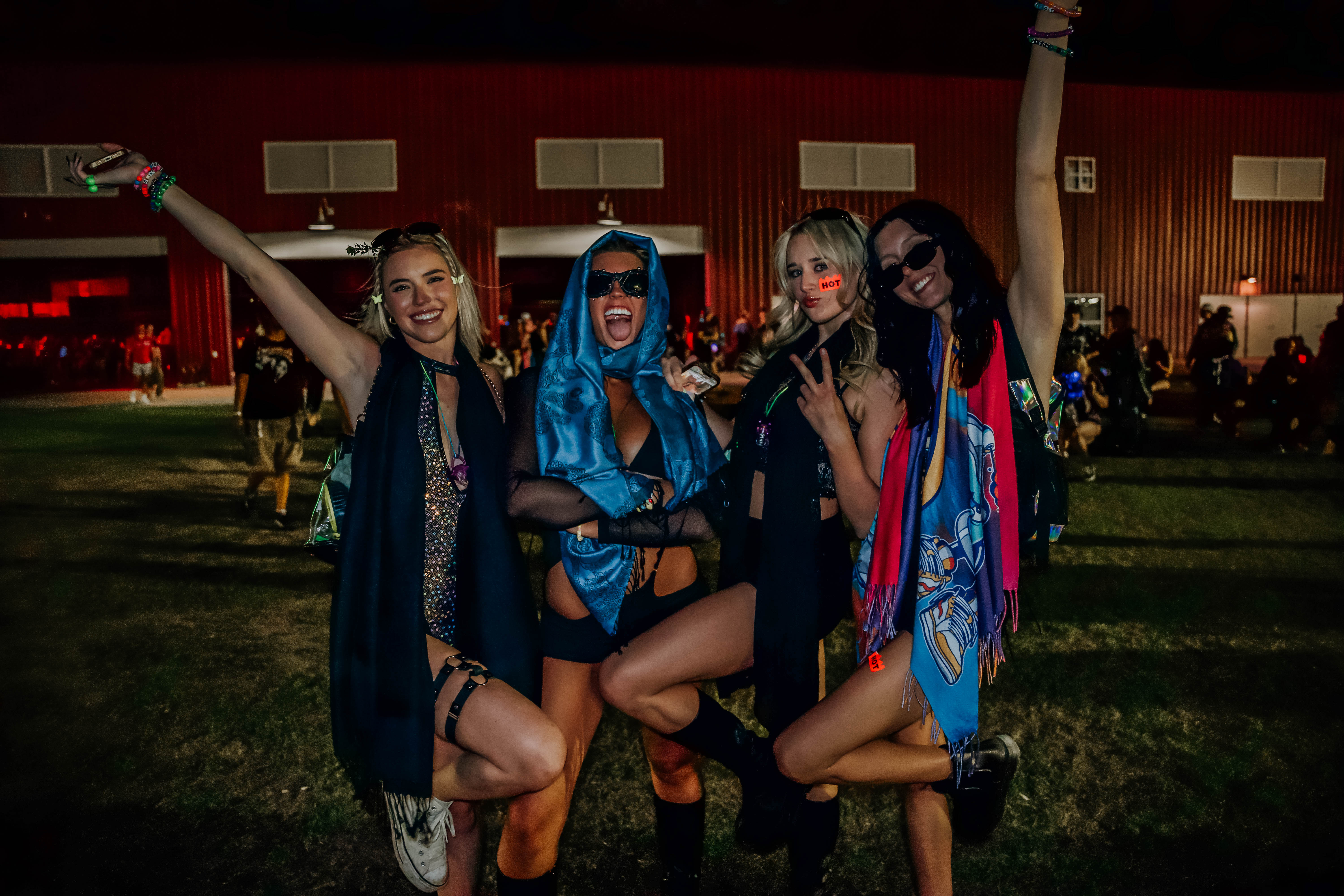 A group of four women posing together at a festival, dressed in colorful outfits with accessories, and expressing excitement and joy.