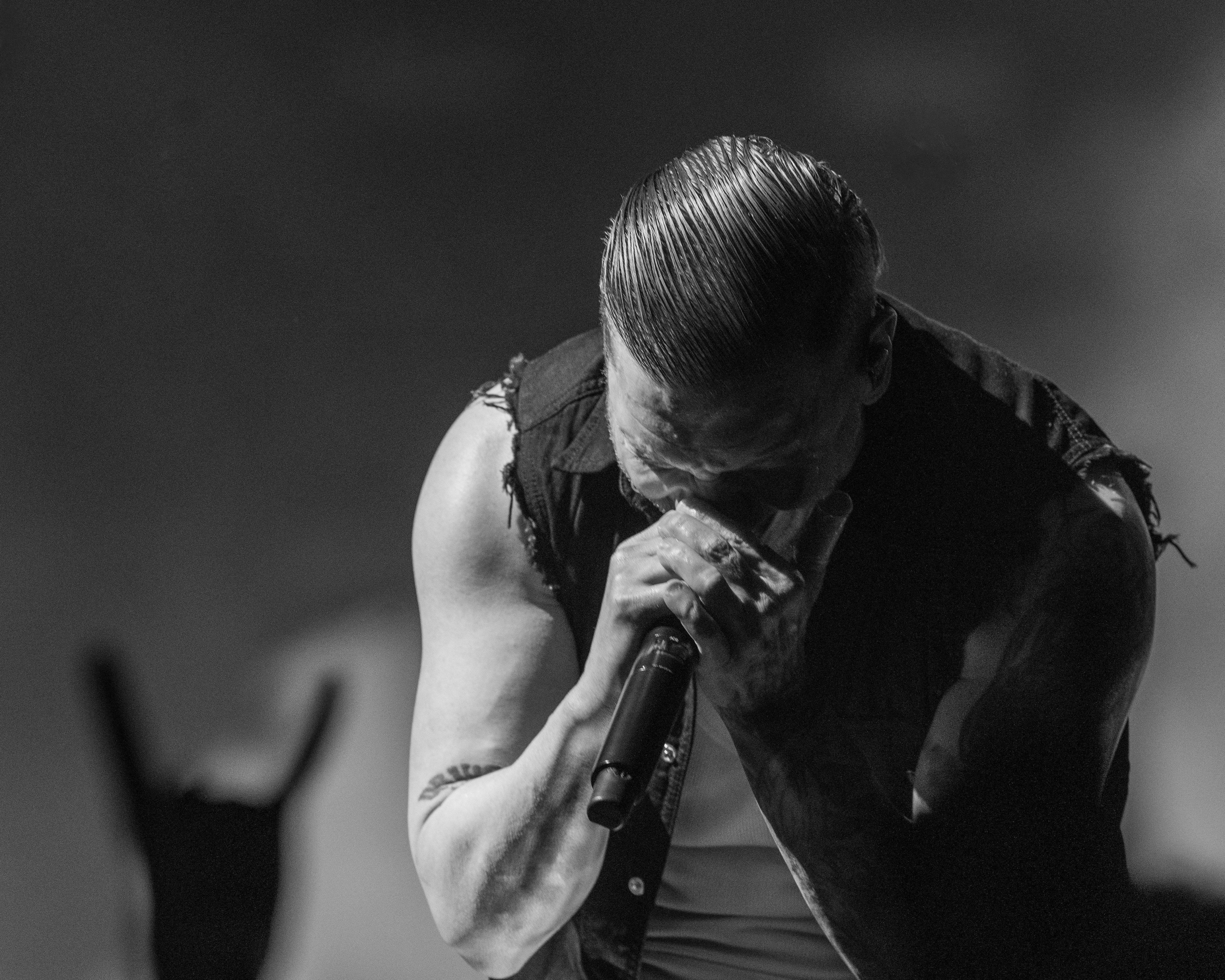 Black and white image of a male vocalist passionately singing into a microphone, with his face partially obscured and a hand gesture from a fan in the foreground.