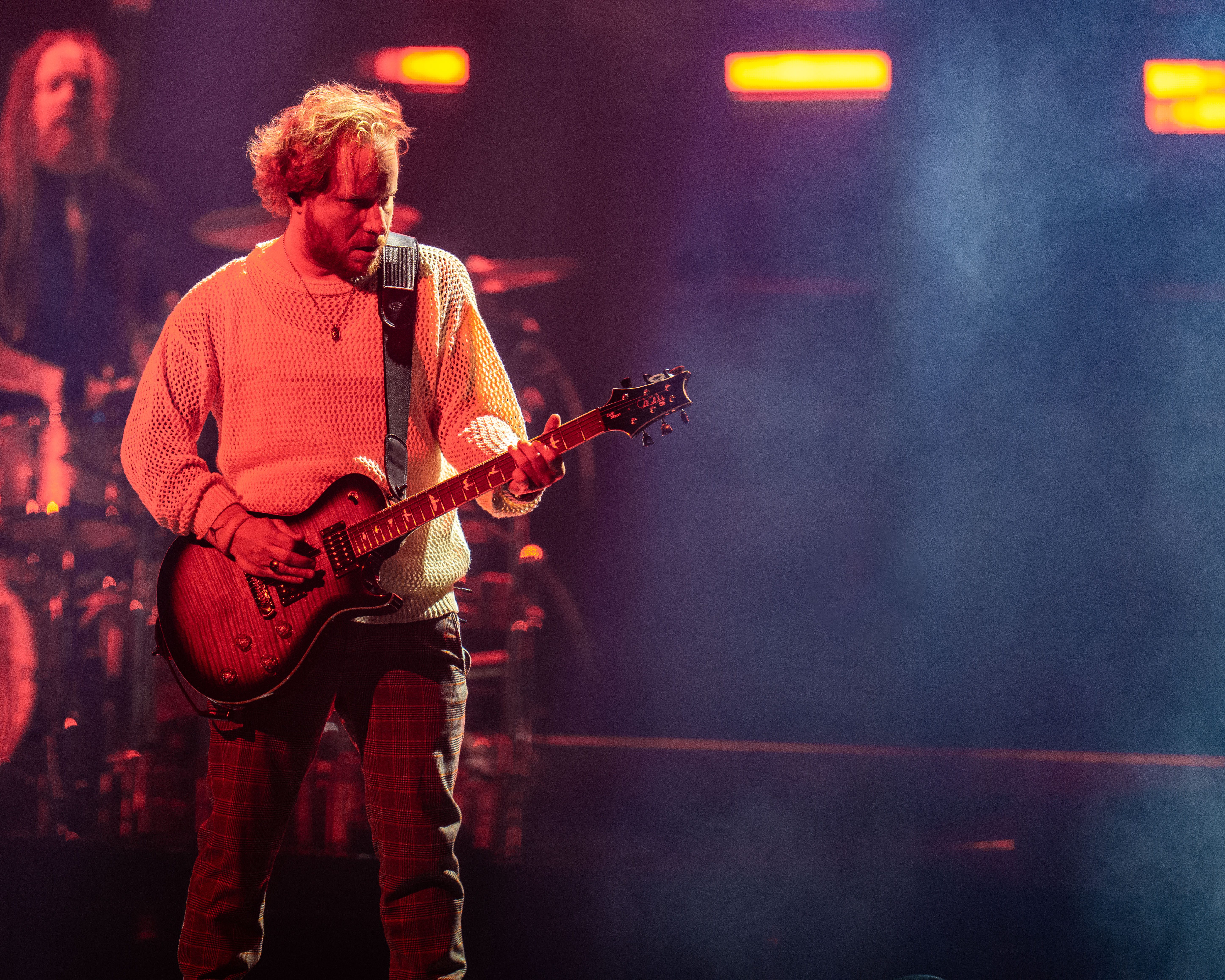 A musician performing on stage with a guitar, wearing a white knitted sweater, surrounded by colorful stage lighting and smoke.
