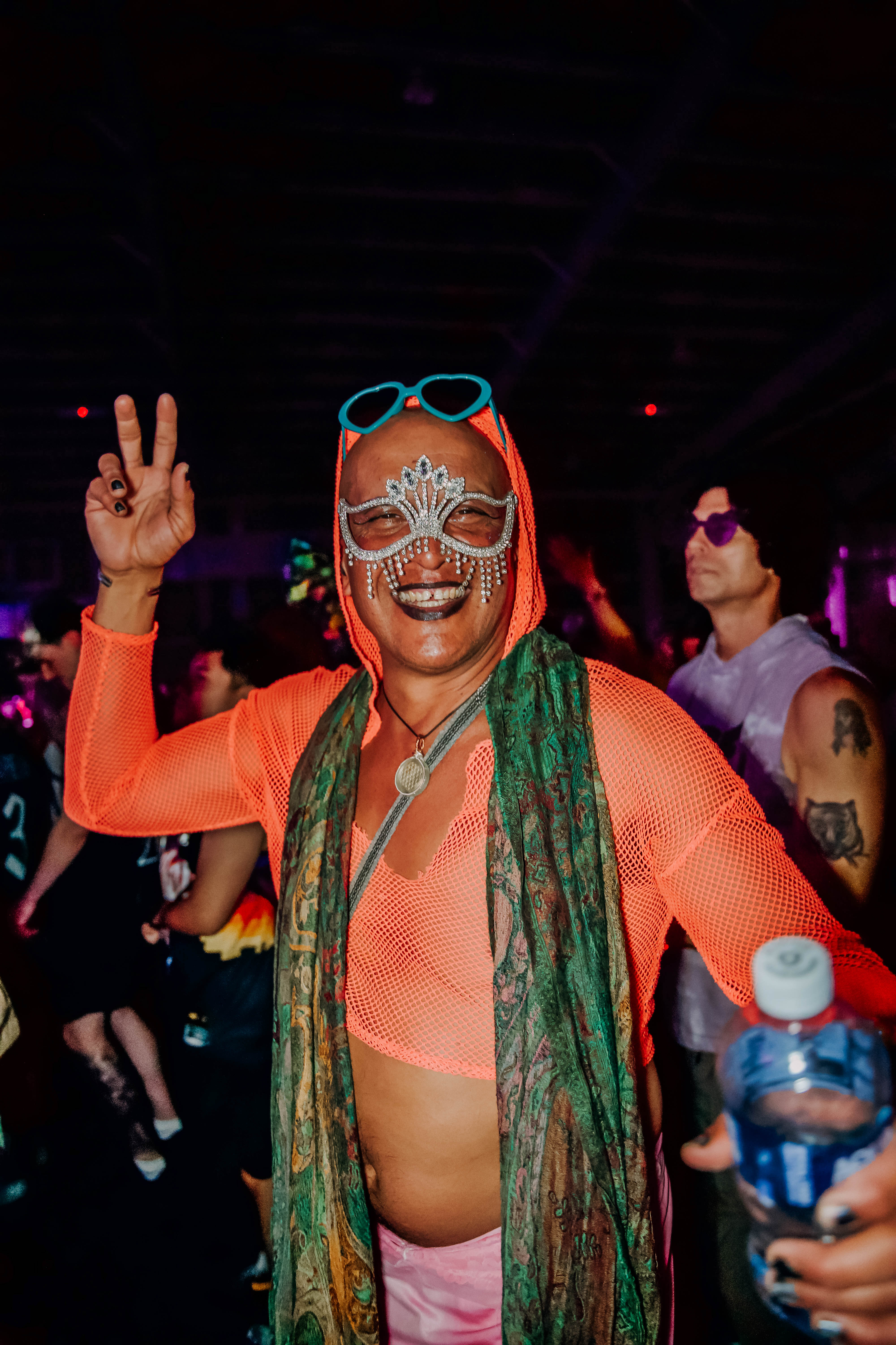 A person wearing a vibrant orange mesh top, colorful sunglasses, and a sparkling masquerade mask poses with a peace sign at a lively event, surrounded by other attendees in a festive atmosphere.