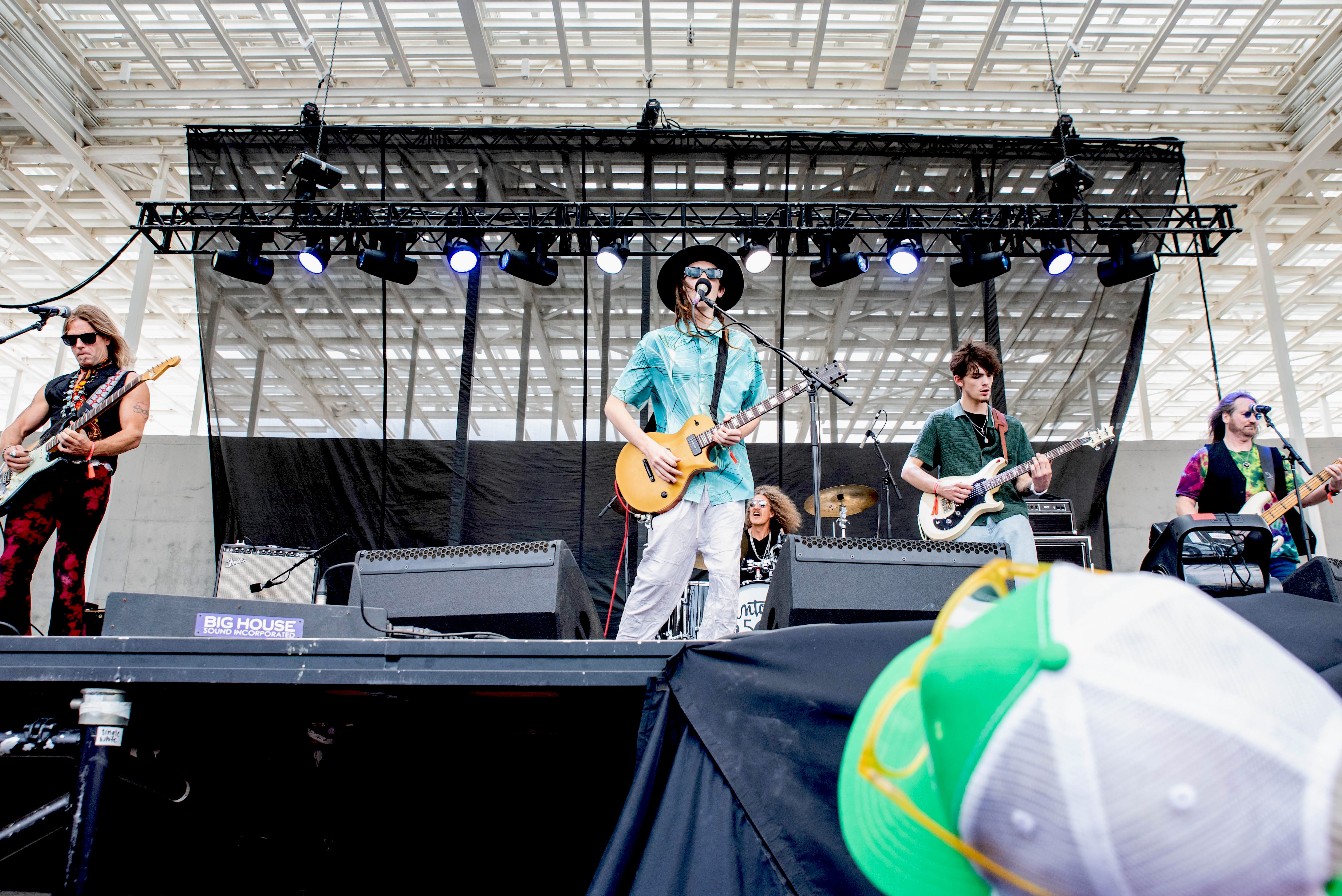 A band performing on stage at the Moody Amphitheater, featuring multiple musicians playing guitars and drums, with a bright, open roof design overhead.