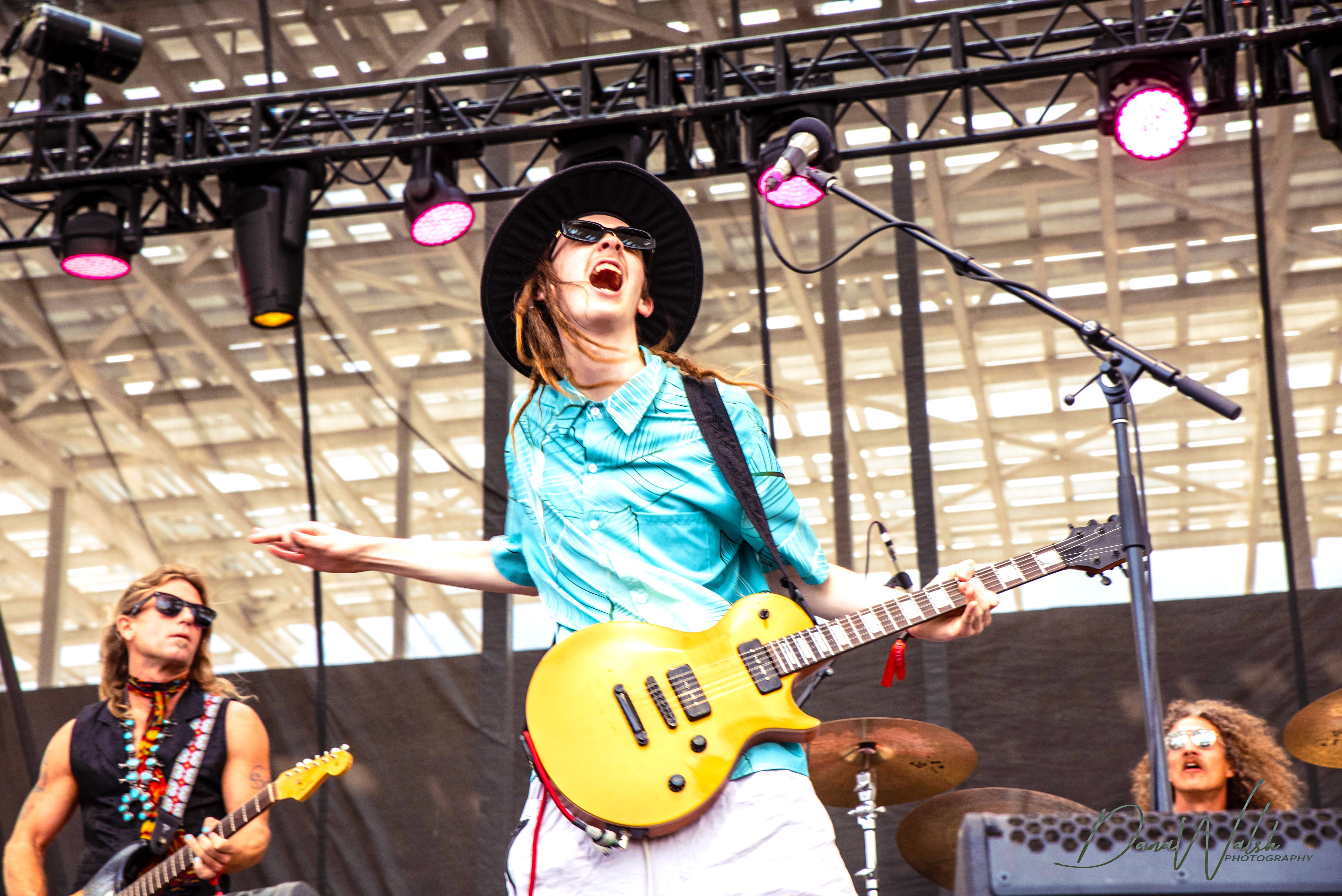 Musician passionately performing on stage with a yellow guitar, wearing sunglasses and a wide-brimmed hat, while a drummer plays in the background.