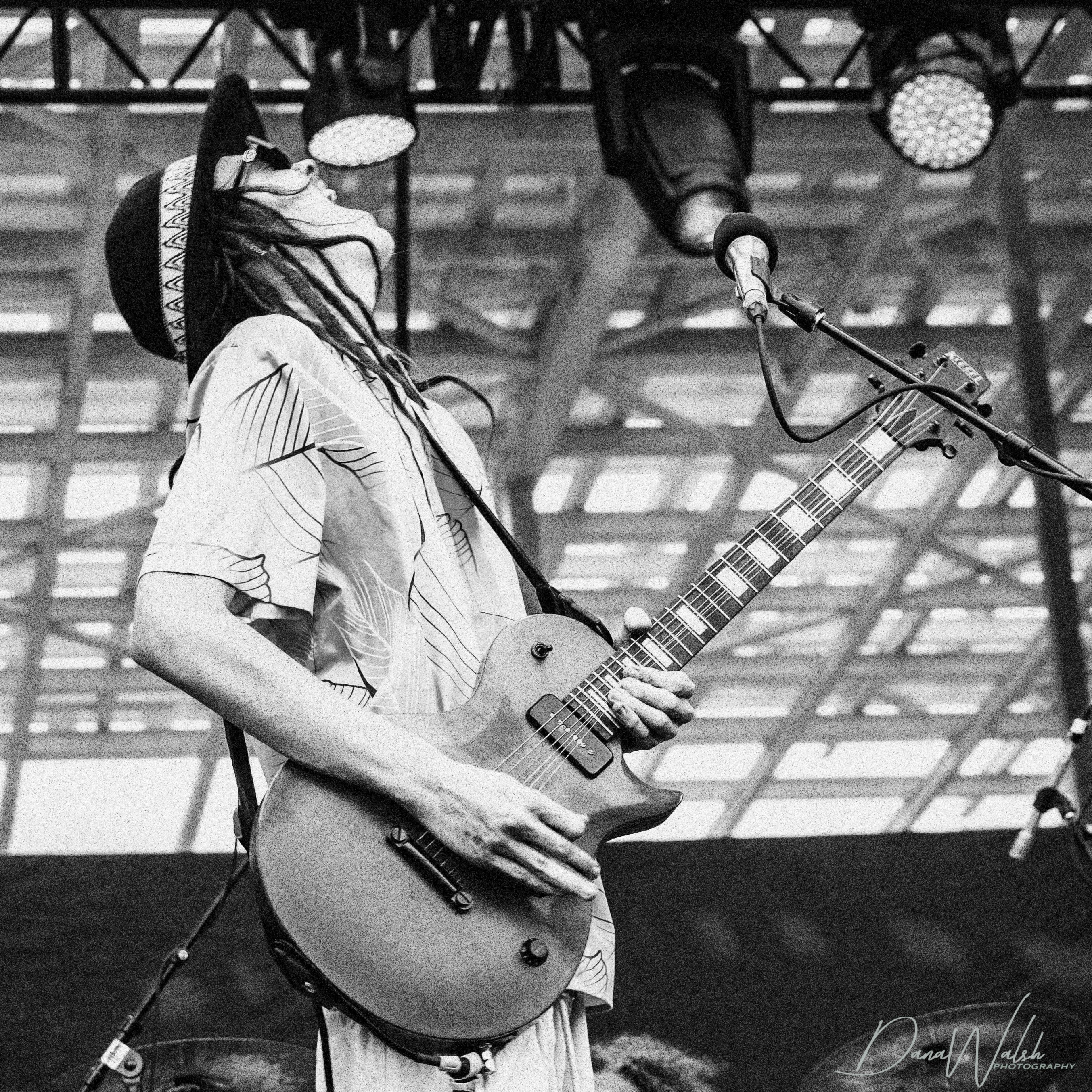 A musician passionately playing an electric guitar on stage, wearing a patterned shirt and a wide-brimmed hat, with a lively atmosphere in the background.