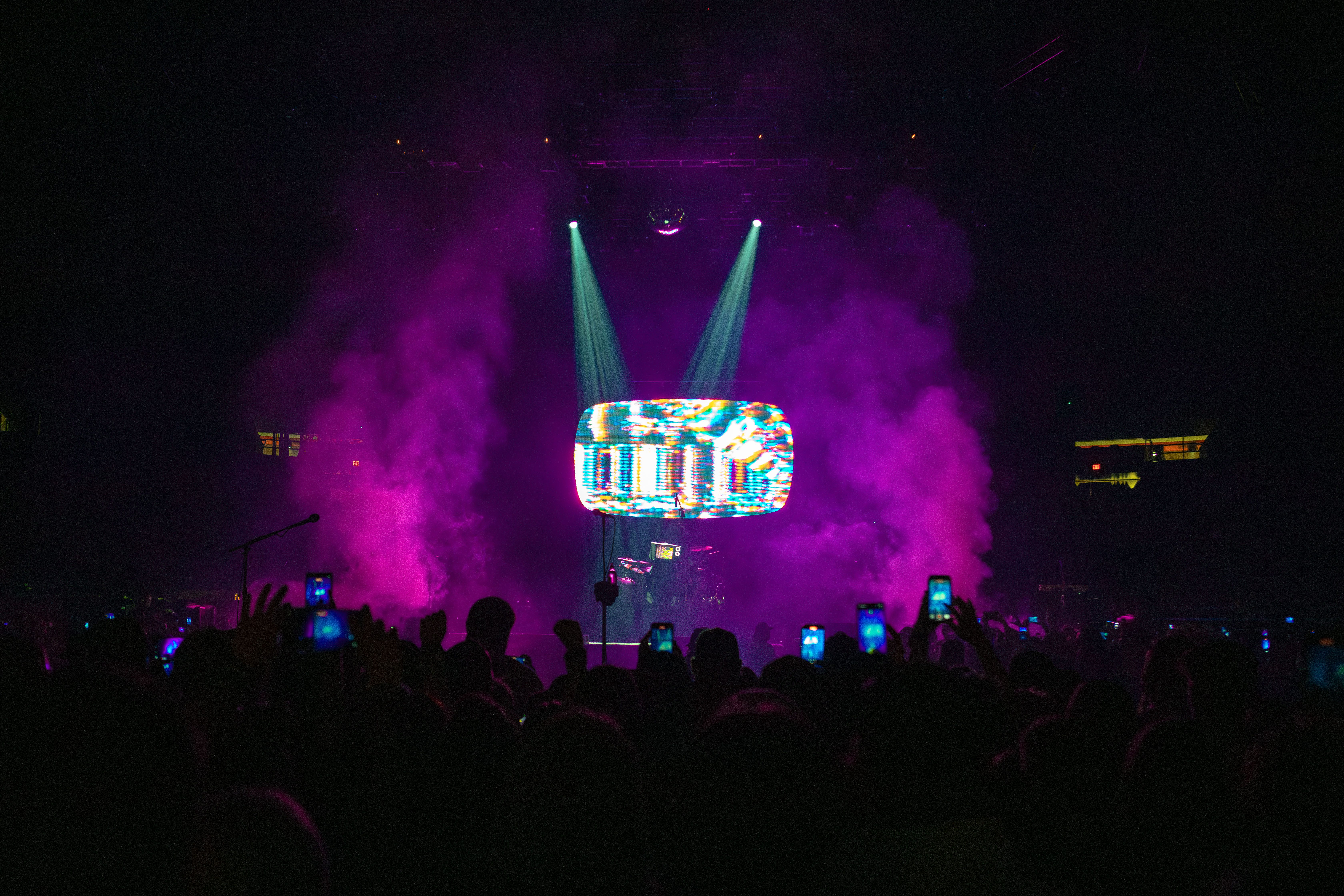 A lively concert scene with vibrant lighting, featuring a performer on stage illuminated by colorful beams and surrounded by a crowd, all holding up phones to capture the moment.
