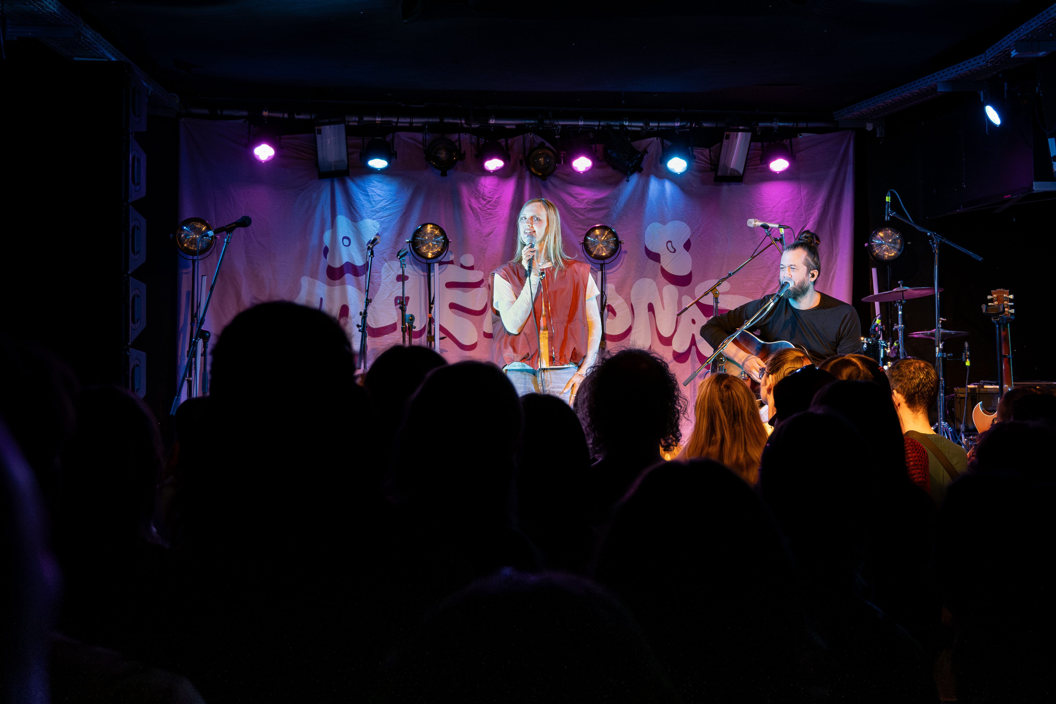 A live music performance featuring two musicians on stage, with a vibrant backdrop and a crowd of fans watching the show.
