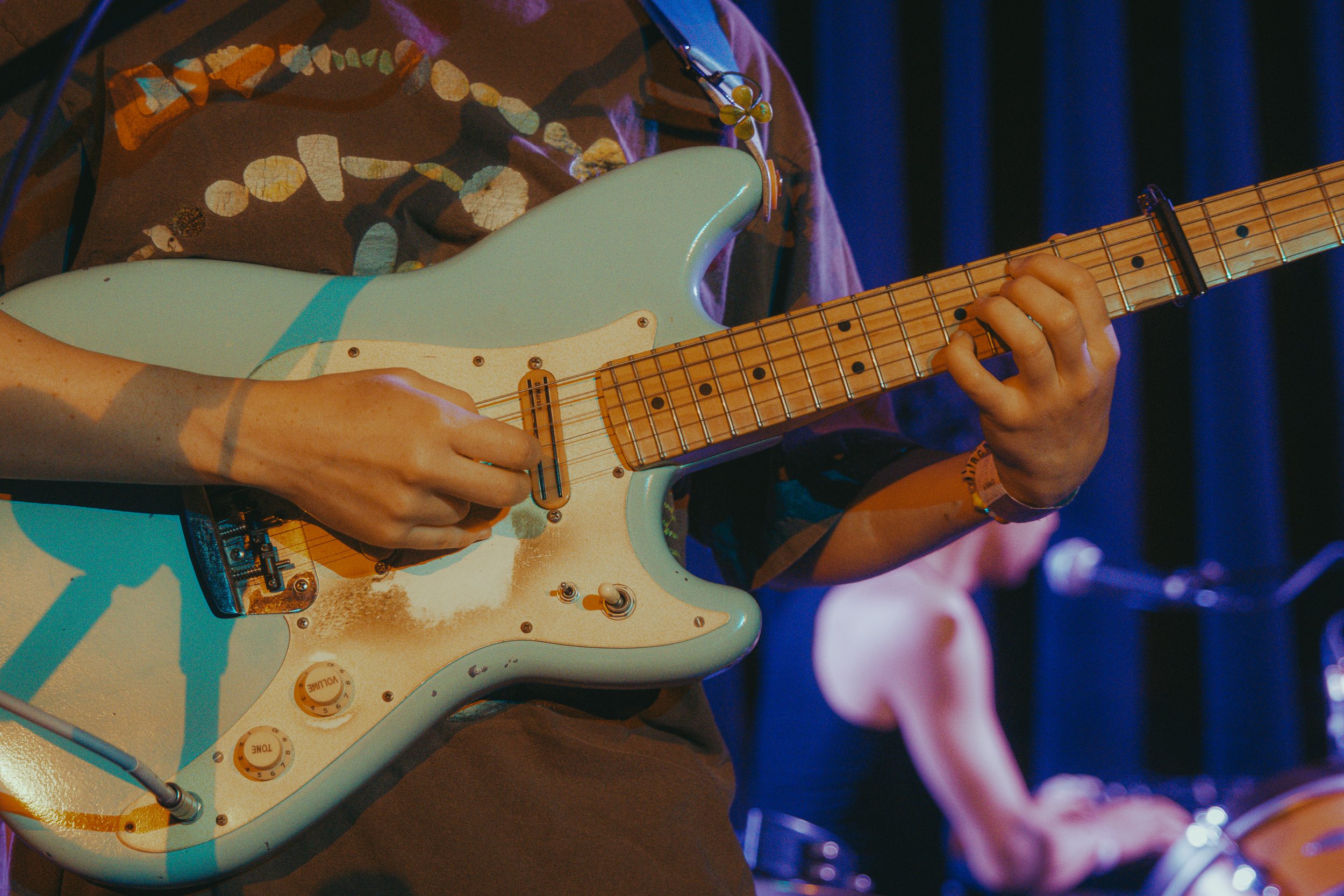 Close-up of a guitarist's hand playing a light blue electric guitar on stage, with blurred band members visible in the background.