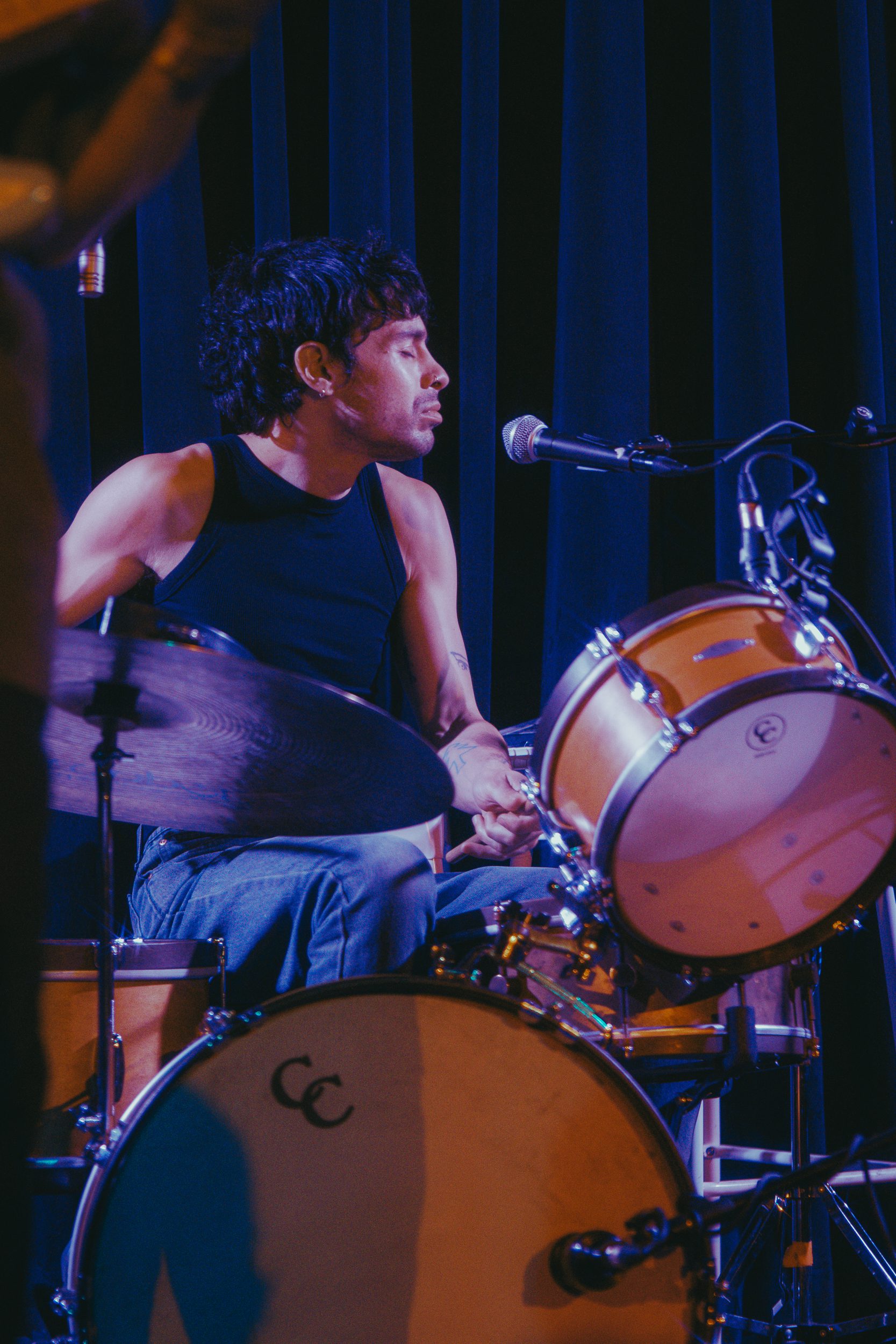 A drummer on stage, focused on playing, with soft lighting and a backdrop of dark curtains.
