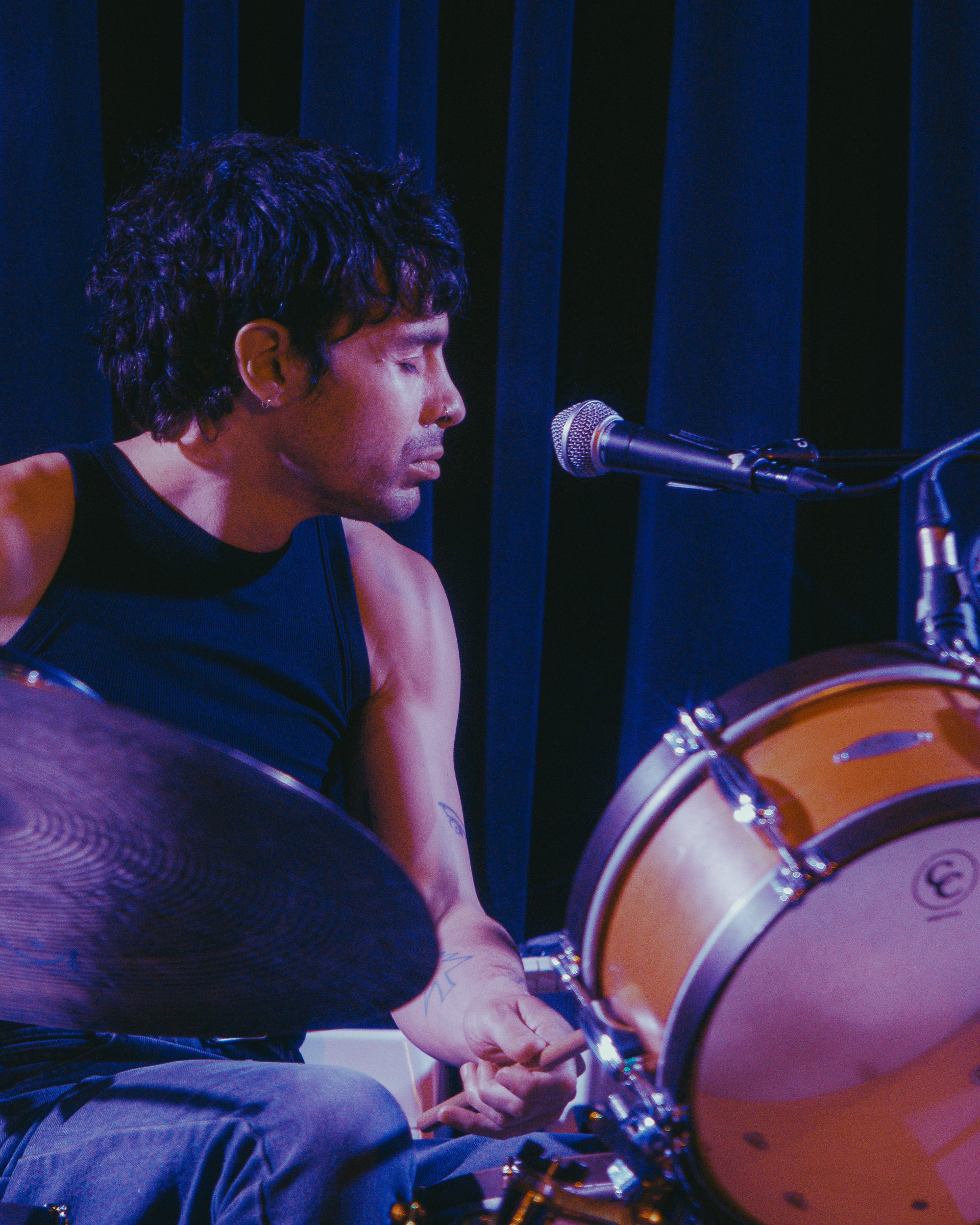 A drummer in a black tank top plays on stage, looking introspective as he grips a drumstick, with softly lit background curtains.