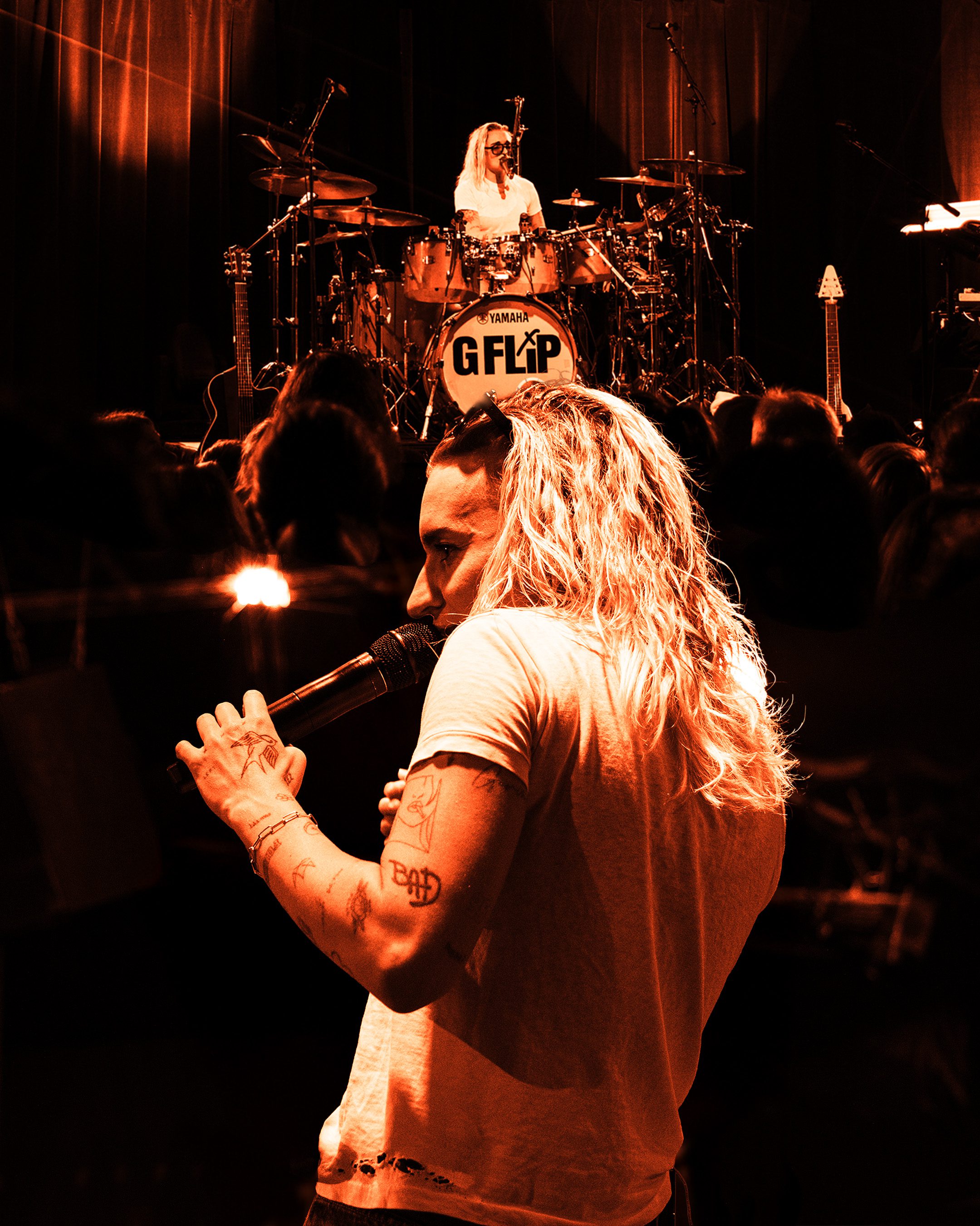 A musician with long, wavy hair stands on stage holding a microphone, while a drummer is visible in the background, creating an energetic performance atmosphere.