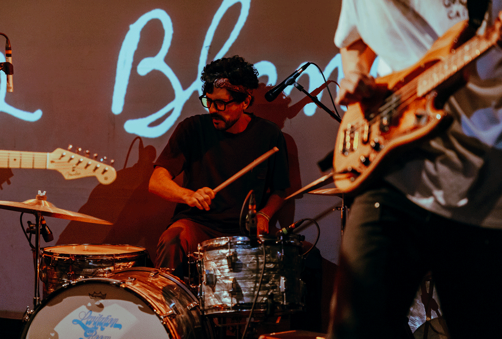 A drummer performing on stage with a confident expression, surrounded by instruments and a colorful backdrop.